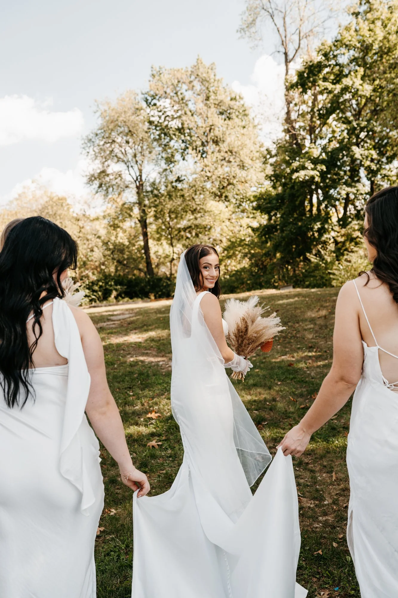 A bride with dark hair, wearing a white dress and veil, holding a bouquet of dried flowers, smiling and walking outdoors with bridesmaids in white dresses on a sunny day with trees in the background.