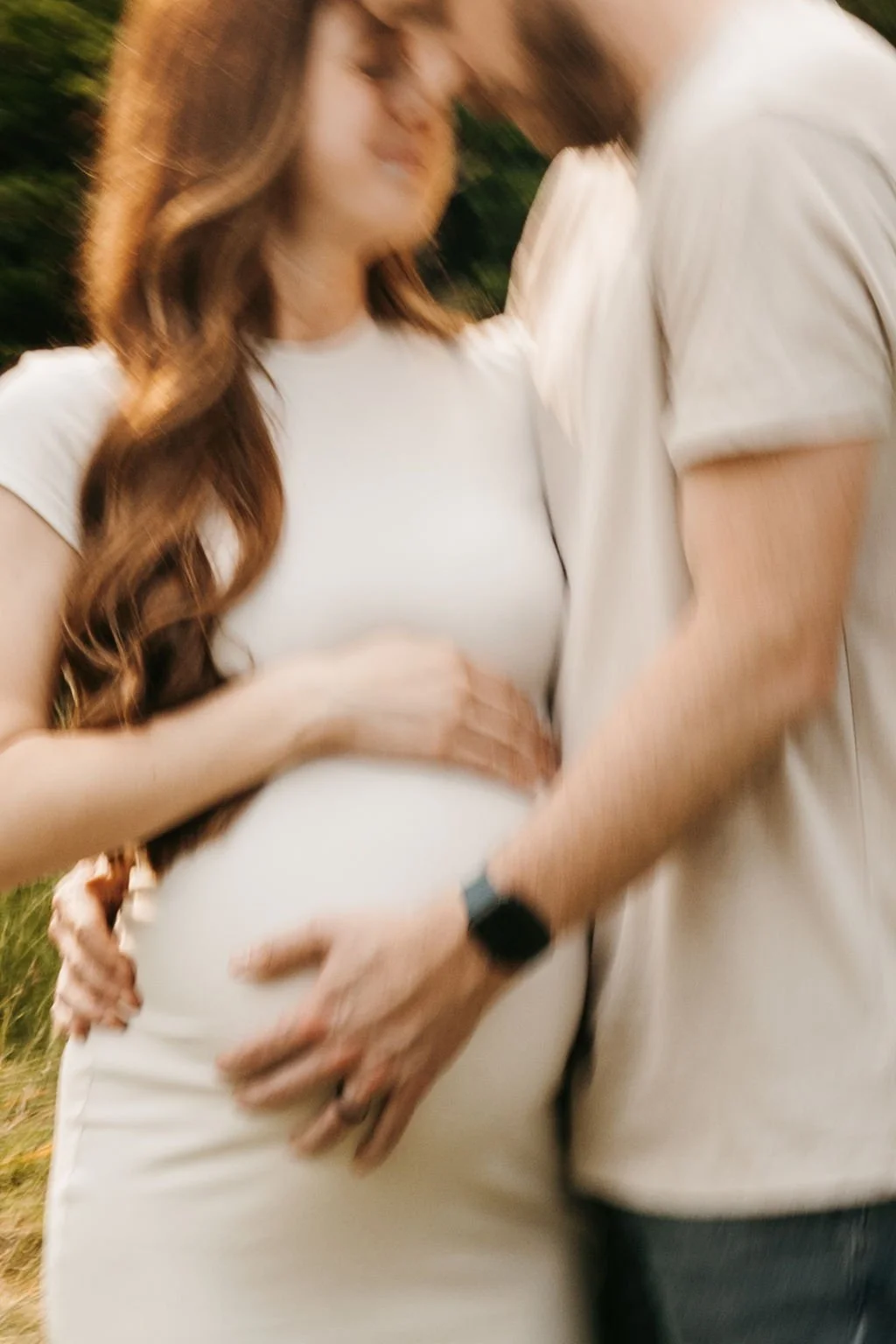 A pregnant woman and a man are outside, holding her belly and kissing each other.