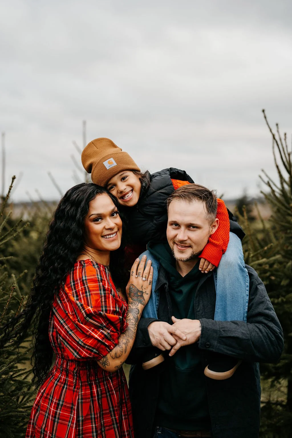A family of three in a Christmas tree farm, with evergreen trees around them. The woman has long curly black hair, tattoos on her arm, and is wearing a red plaid dress. The man has short light brown hair, light facial hair, and is wearing a black jacket with a green hoodie. The child, wearing a brown beanie and black jacket, is on the man's shoulders, smiling widely.