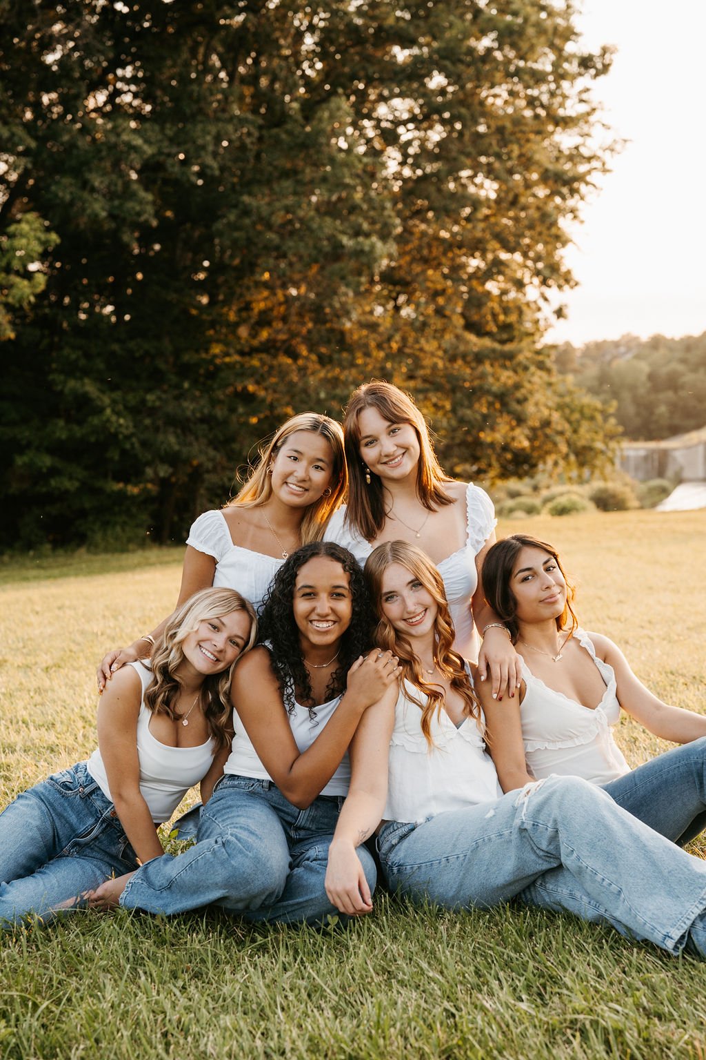 Group of six young women sitting on grass in a park during sunset, smiling and looking at the camera.