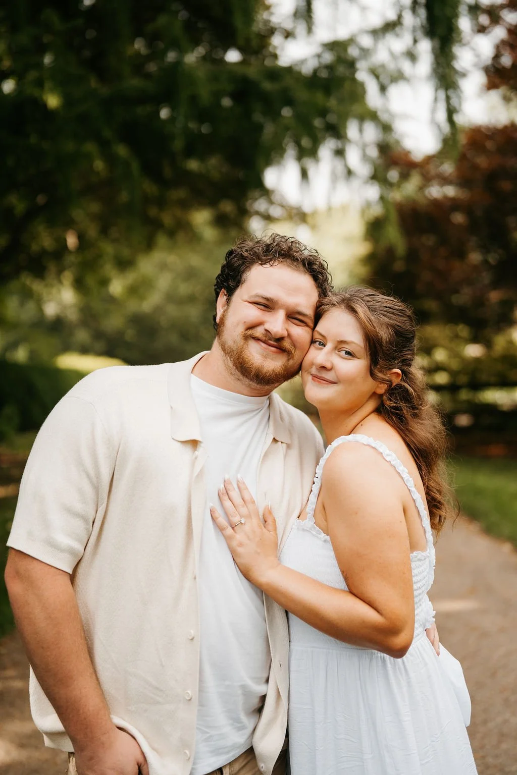 A smiling man and woman embracing outdoors on a sunny day, surrounded by trees.
