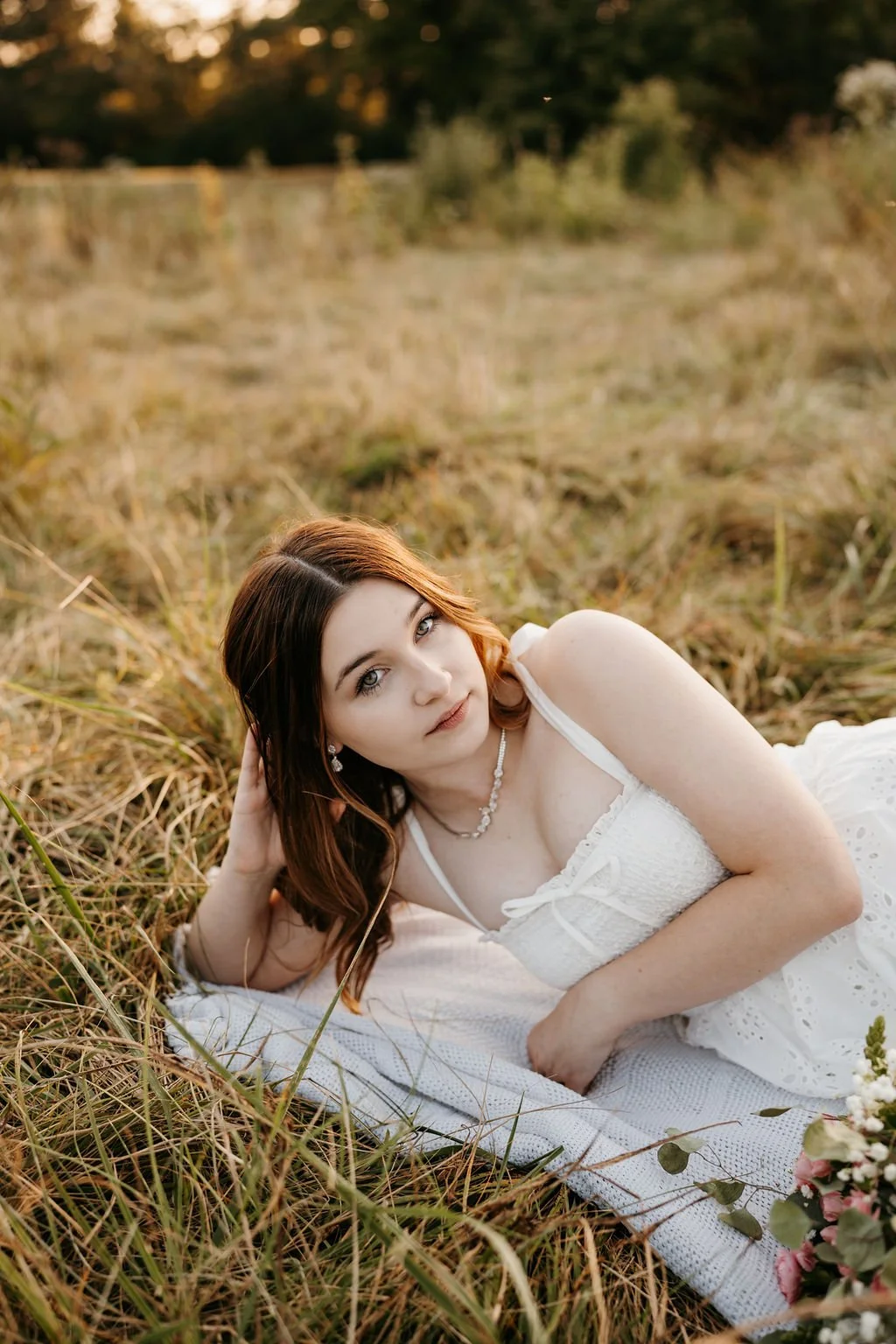 A woman lying on a white blanket in a grassy field during sunset, wearing a white dress and jewelry, and looking directly at the camera.
