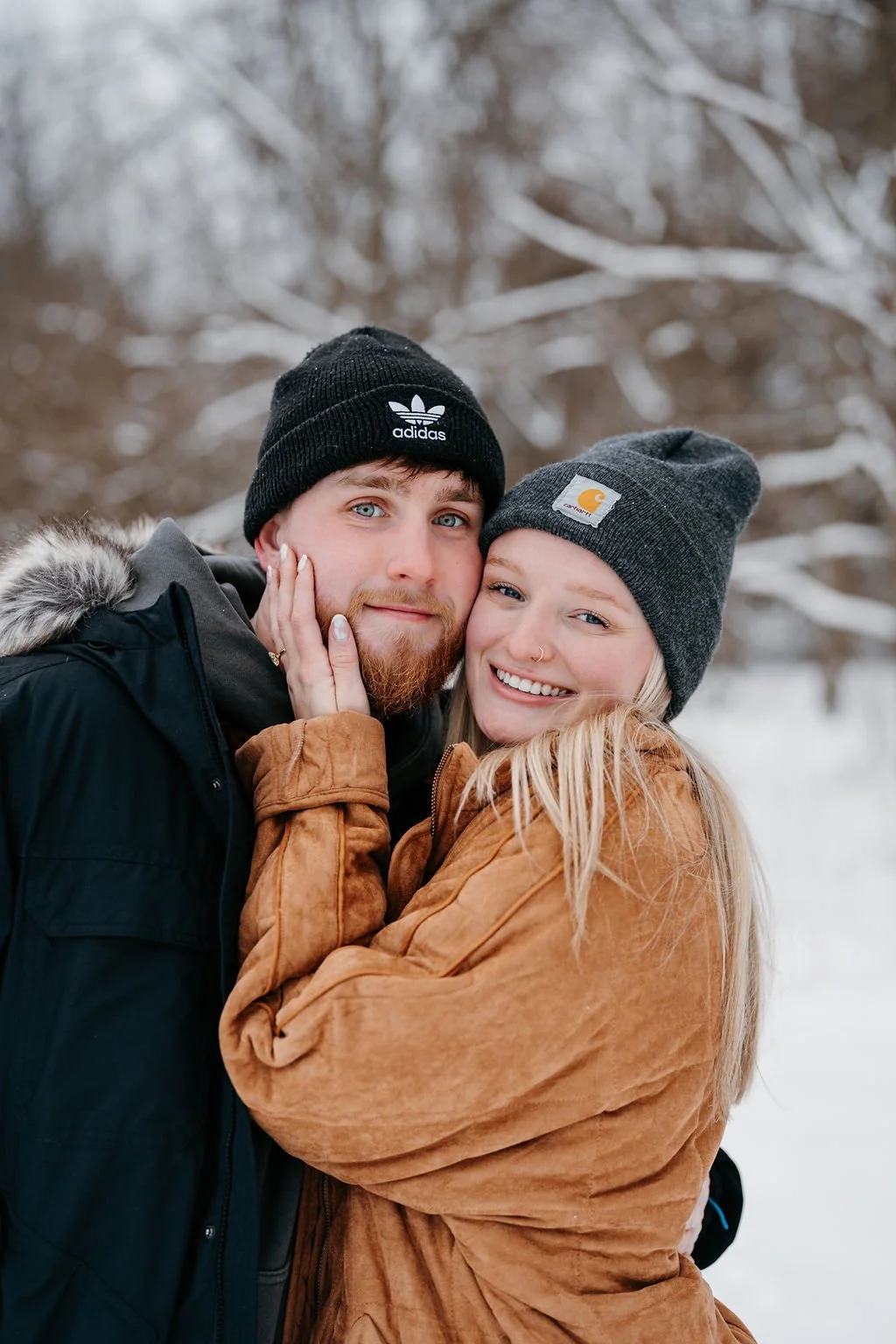 A young couple with light skin embracing outdoors in a snowy winter setting, both wearing winter hats and jackets, smiling at the camera.