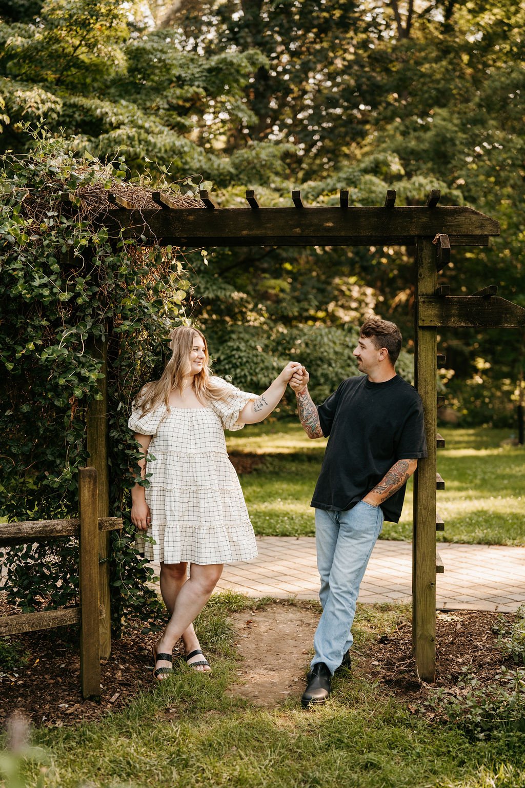 A young woman and man are holding hands and smiling at each other under a wooden garden archway, surrounded by greenery in an outdoor park setting.