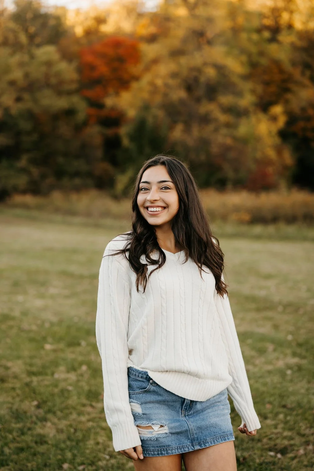 A young woman with long dark hair and light skin, smiling and looking at the camera, standing outdoors in a grassy field with autumn-colored trees in the background. She is wearing a white cable-knit sweater and a distressed denim skirt.