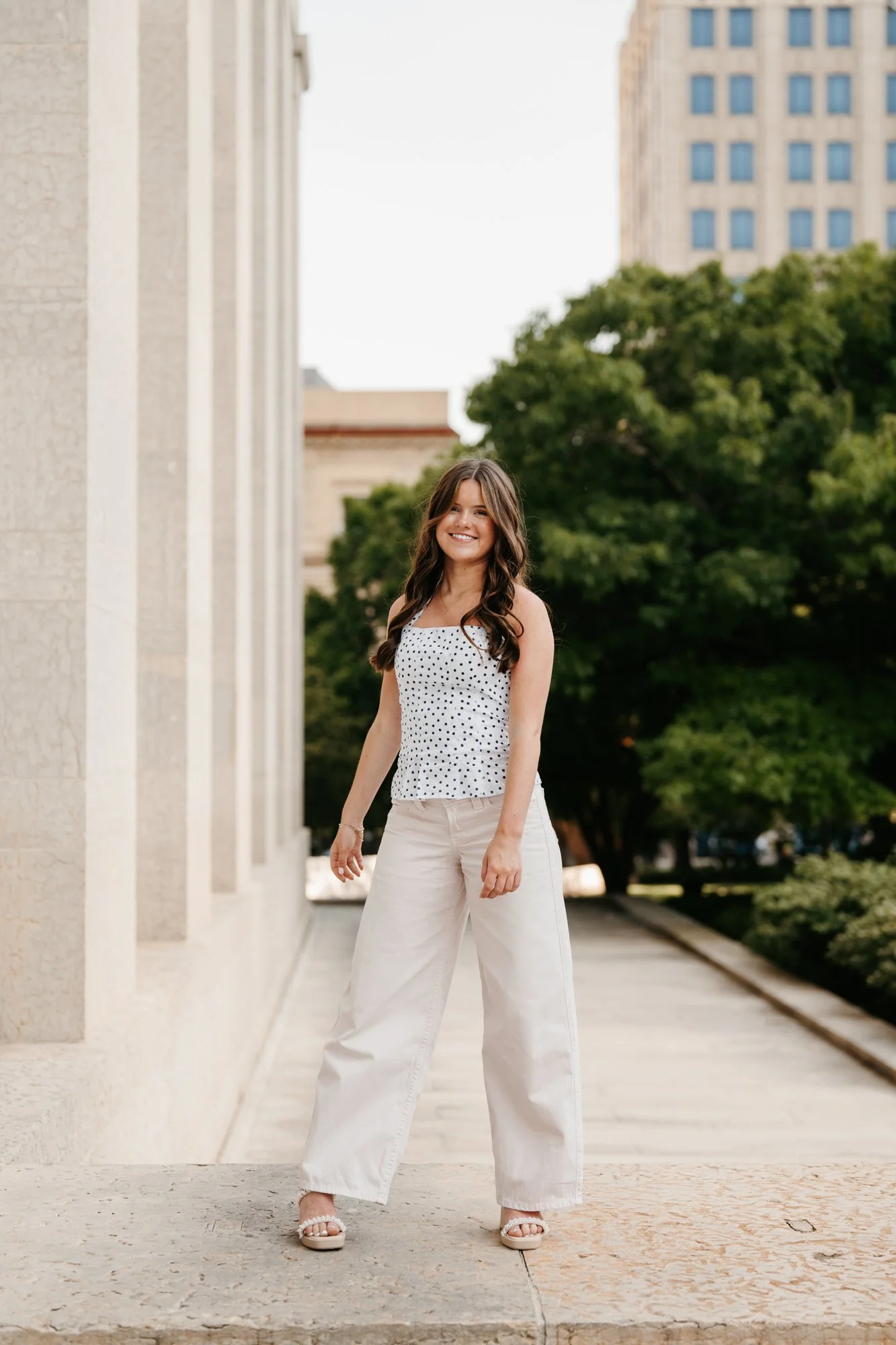 A young woman with long brown hair, wearing a white sleeveless polka dot top, wide-leg beige pants, and beige chunky heels, standing outdoors on a sidewalk with a smile. Green trees and tall buildings are in the background.
