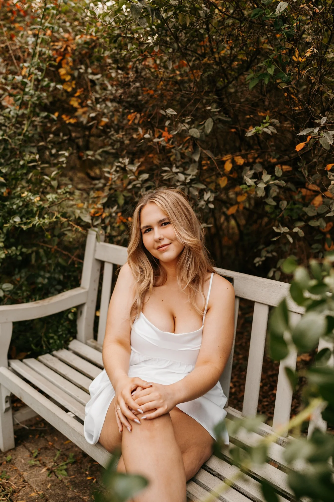 A young woman with wavy blonde hair sitting on a white garden bench in front of lush green foliage and autumn-colored leaves, wearing a white spaghetti strap dress.