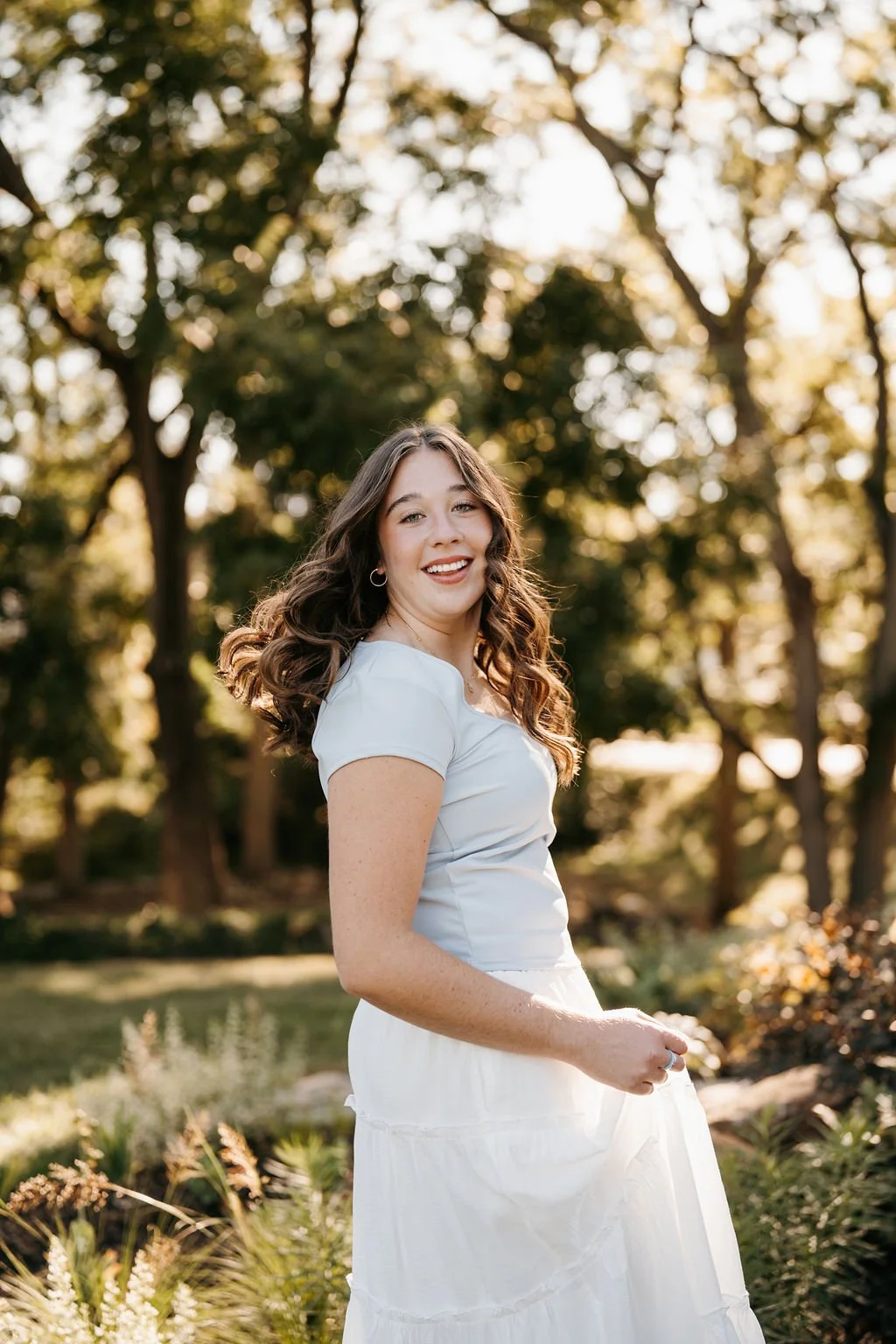 A young woman with long, curly brown hair, wearing a white dress, standing outdoors in a wooded area during golden hour, smiling and looking at the camera.
