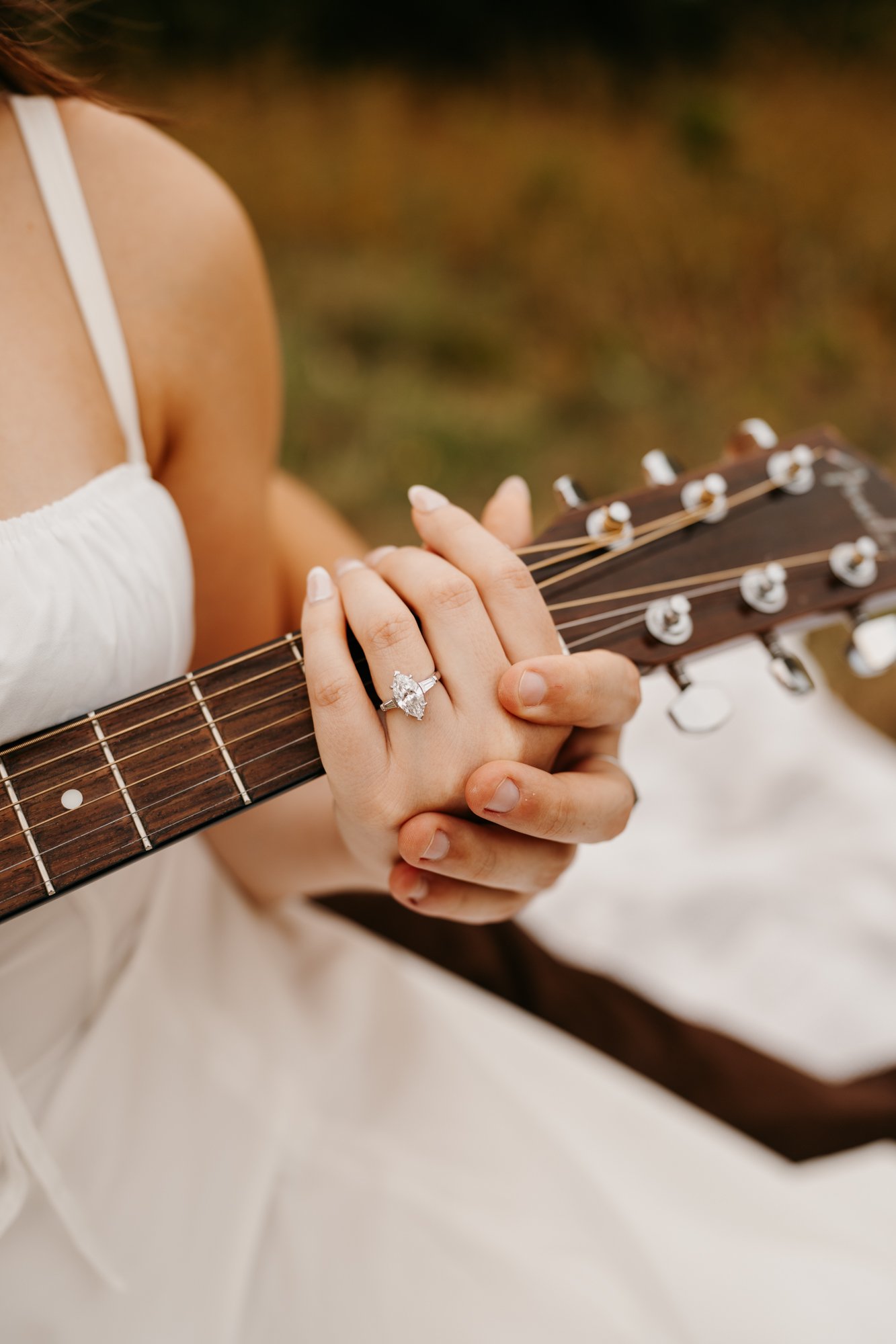 A woman wearing a white dress holding a guitar, with her left hand resting on the neck, showing a diamond engagement ring.