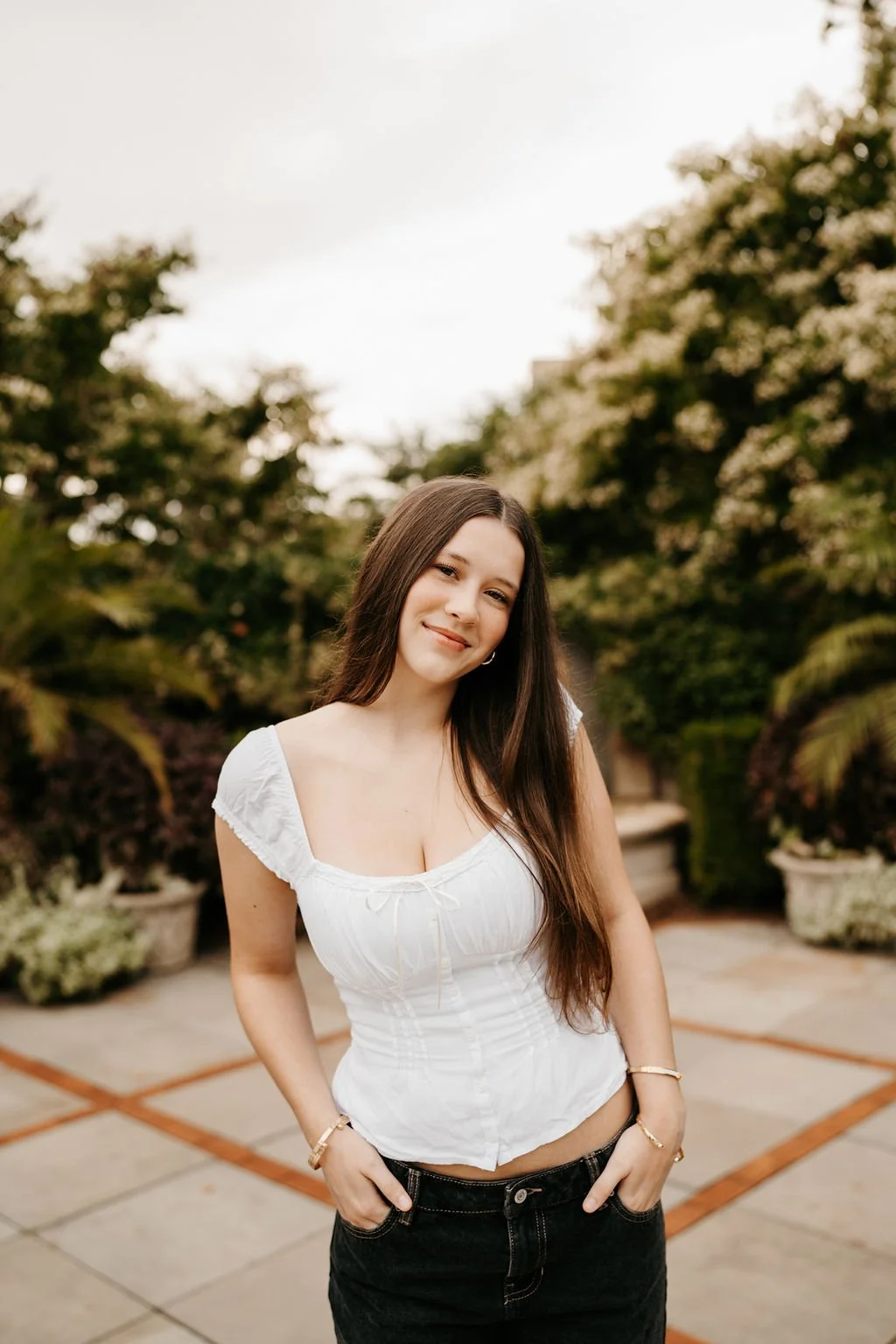 Young woman with long brown hair wearing a white top and black jeans standing outdoors with greenery and potted plants in the background.
