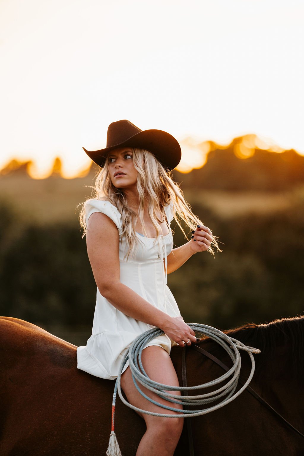 Woman with long blonde hair wearing a brownie hat and white dress, riding a horse at sunset, holding a lasso.