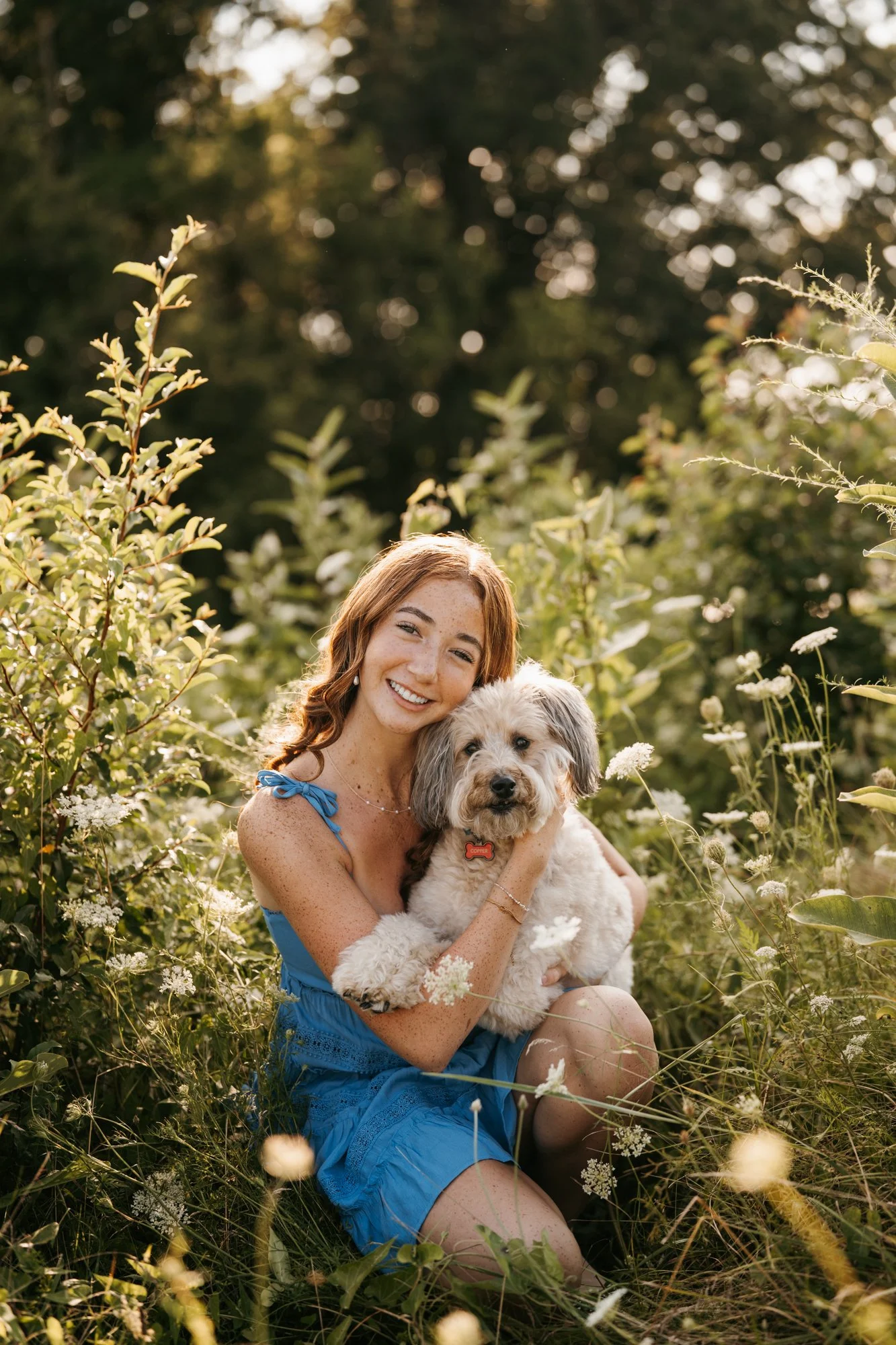A young woman with red hair and freckles wearing a blue dress, sitting in a field of tall grass and wildflowers, smiling while holding a fluffy dog with grey and white fur, during golden hour.