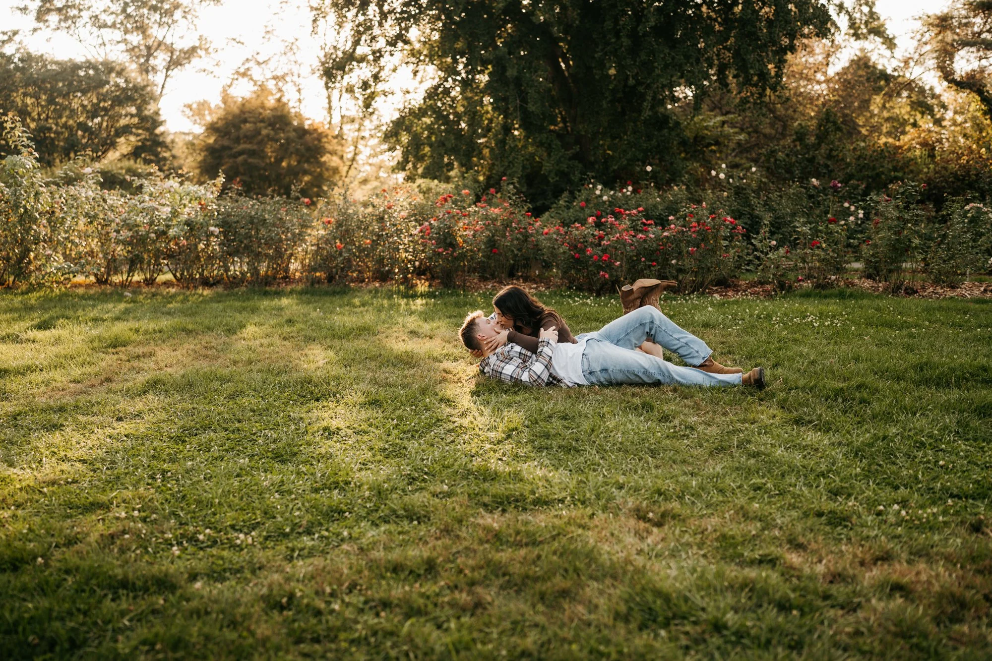 A couple lying on the grass, kissing, in a park during sunset, surrounded by trees and blooming flowers.