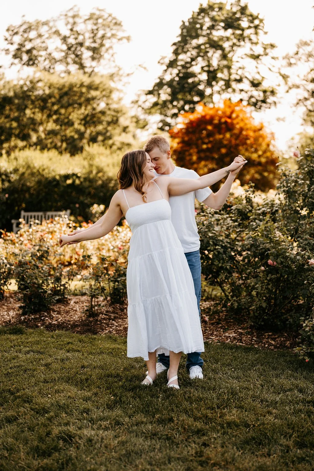 A couple dancing outdoors in a garden with bushes and trees in the background, during late afternoon or early evening, with warm lighting.