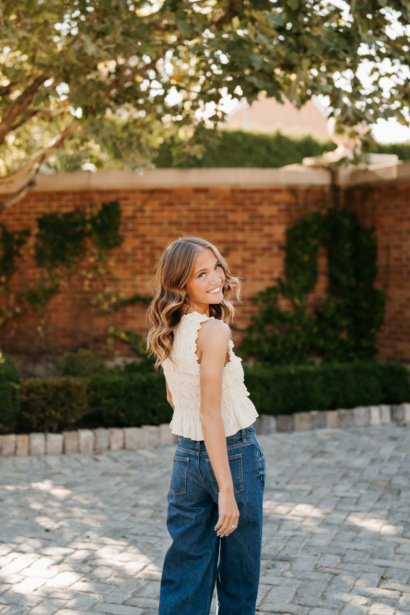 A woman with wavy brown hair wearing a cream-colored sleeveless top and blue jeans, smiling outdoors on a sunny day with a brick wall and greenery in the background.
