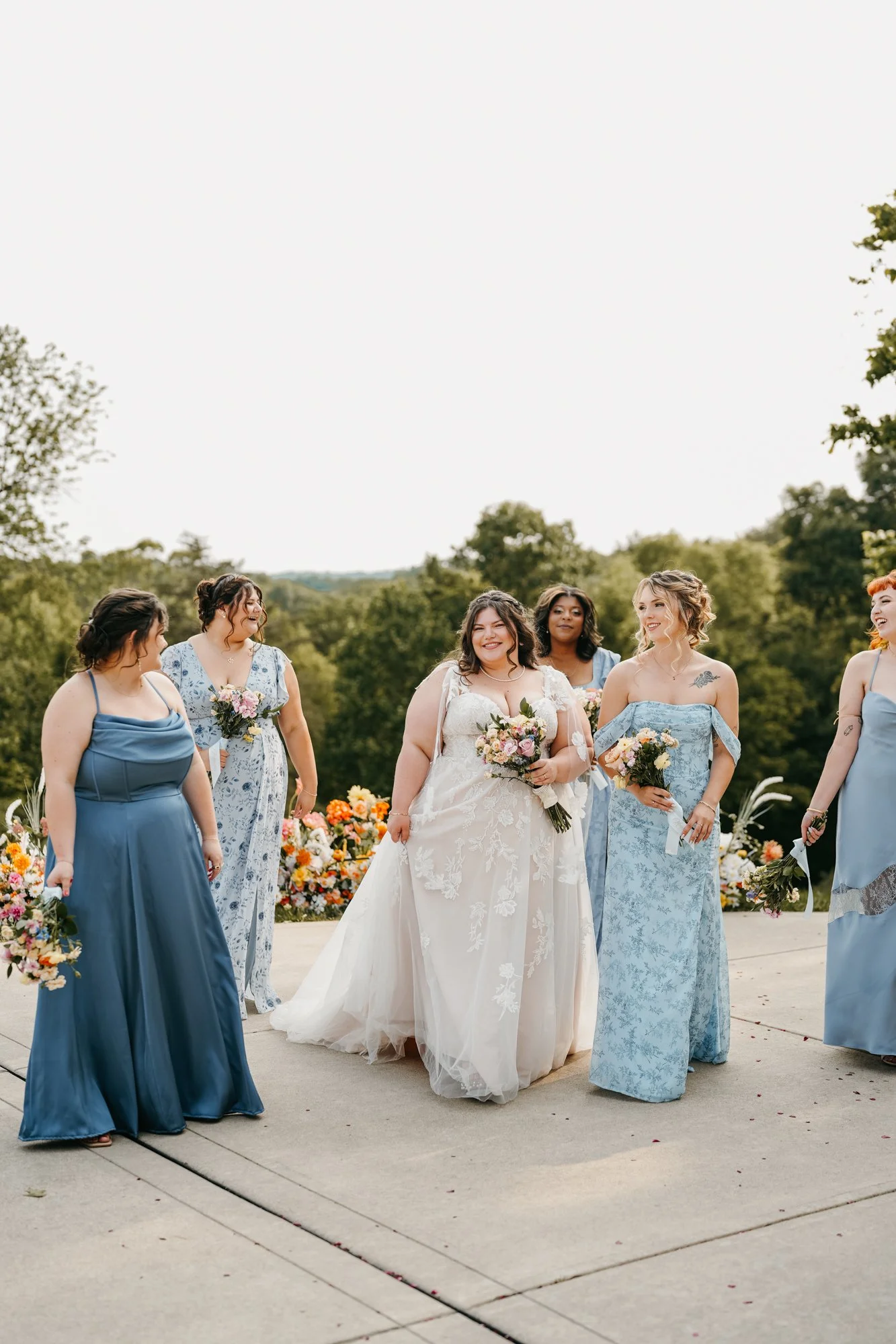 A group of women, including a bride in a white wedding dress, outside during daytime. They are holding bouquets and wearing various shades of blue dresses, standing on a concrete surface with floral arrangements behind them and greenery in the backgr