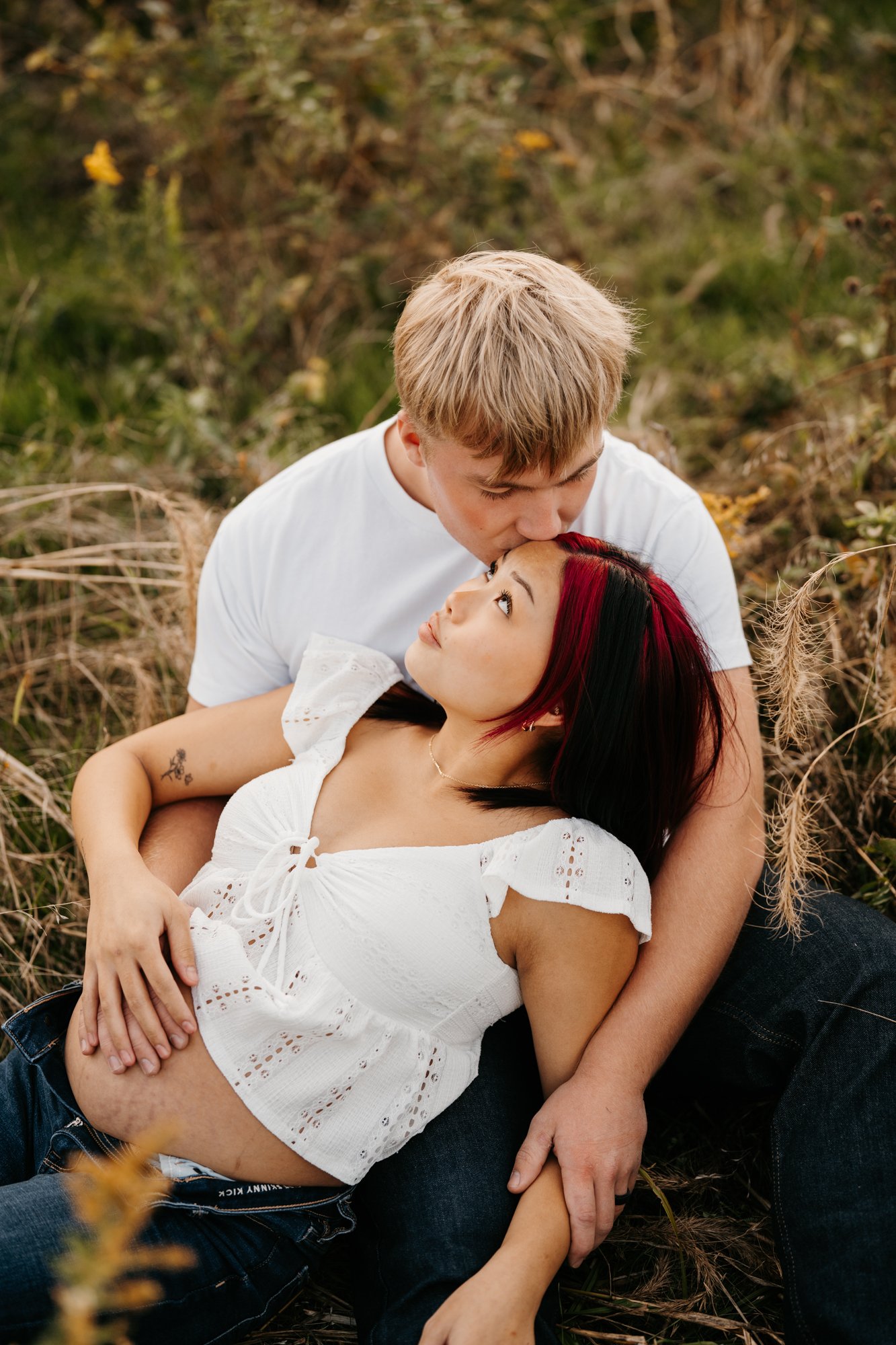 A young couple in an outdoor setting, with the man kissing the woman's forehead while she lies in his lap, surrounded by tall grass and natural foliage.
