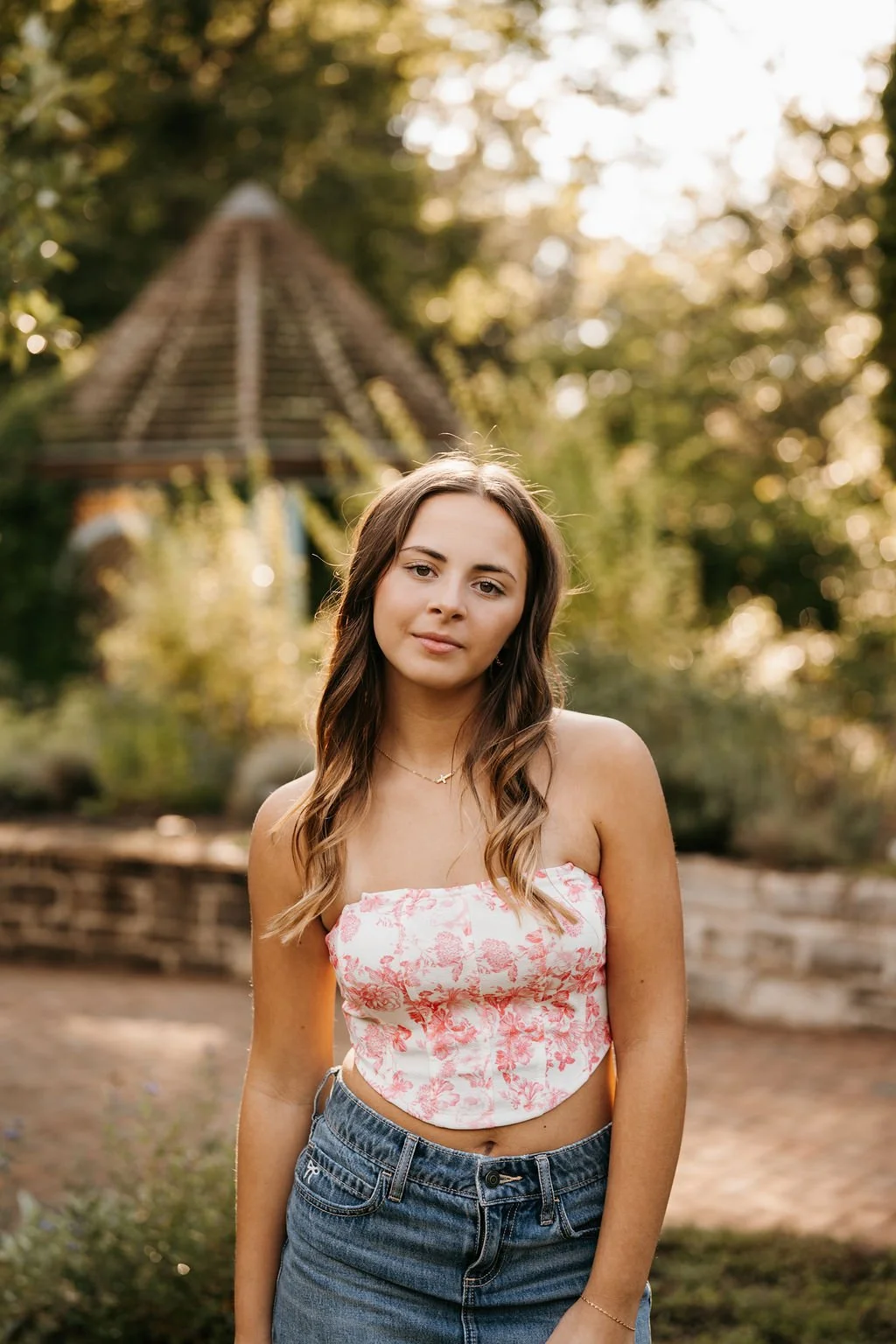 Young woman with long wavy brown hair in a pink and white floral crop top and blue jeans, standing outdoors with trees and a small wooden structure in the background during golden hour.