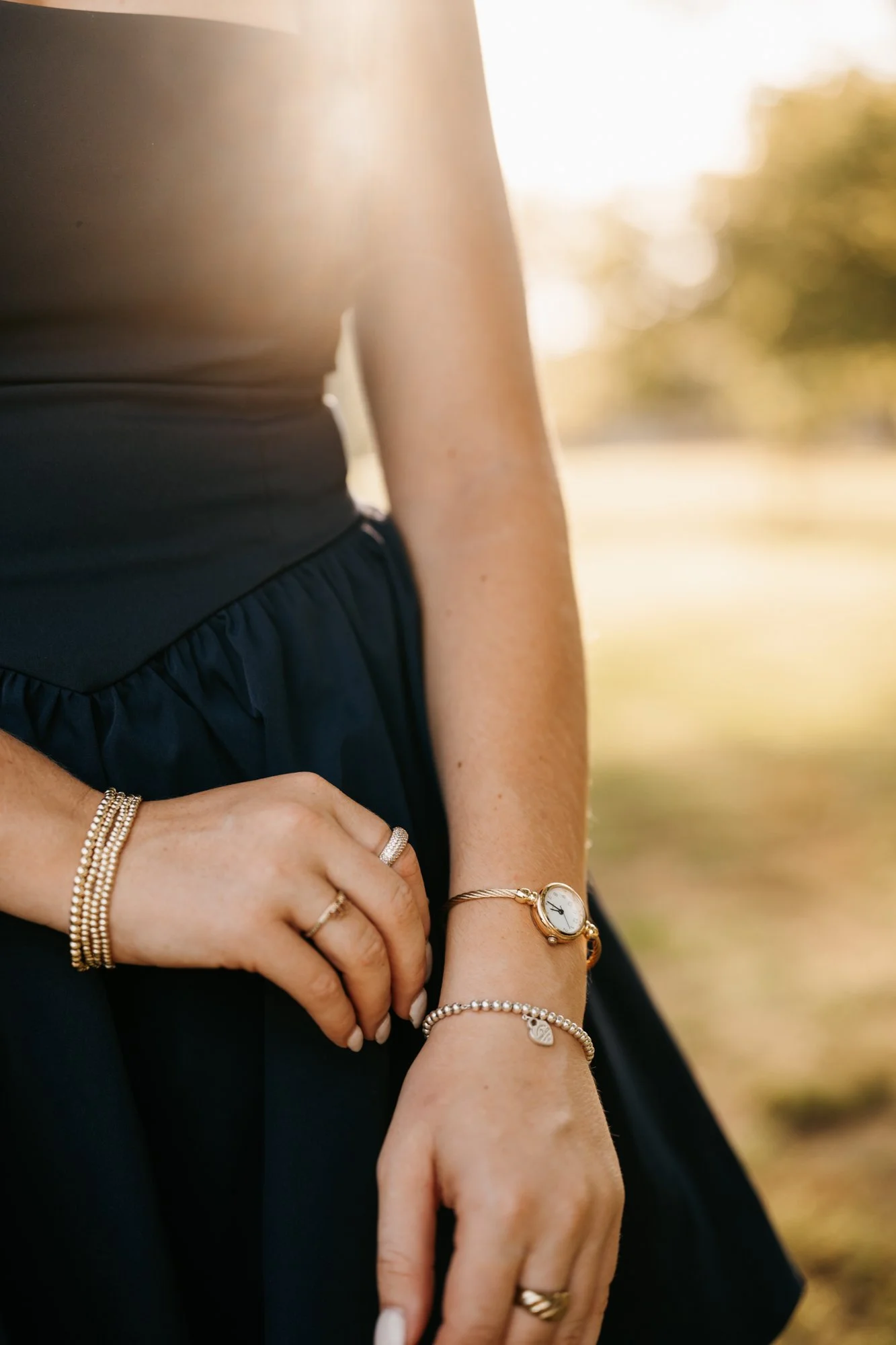 Close-up of a woman's hand and wrist adorned with jewelry, including bracelets and rings, against a blurred outdoor background with sunlight.