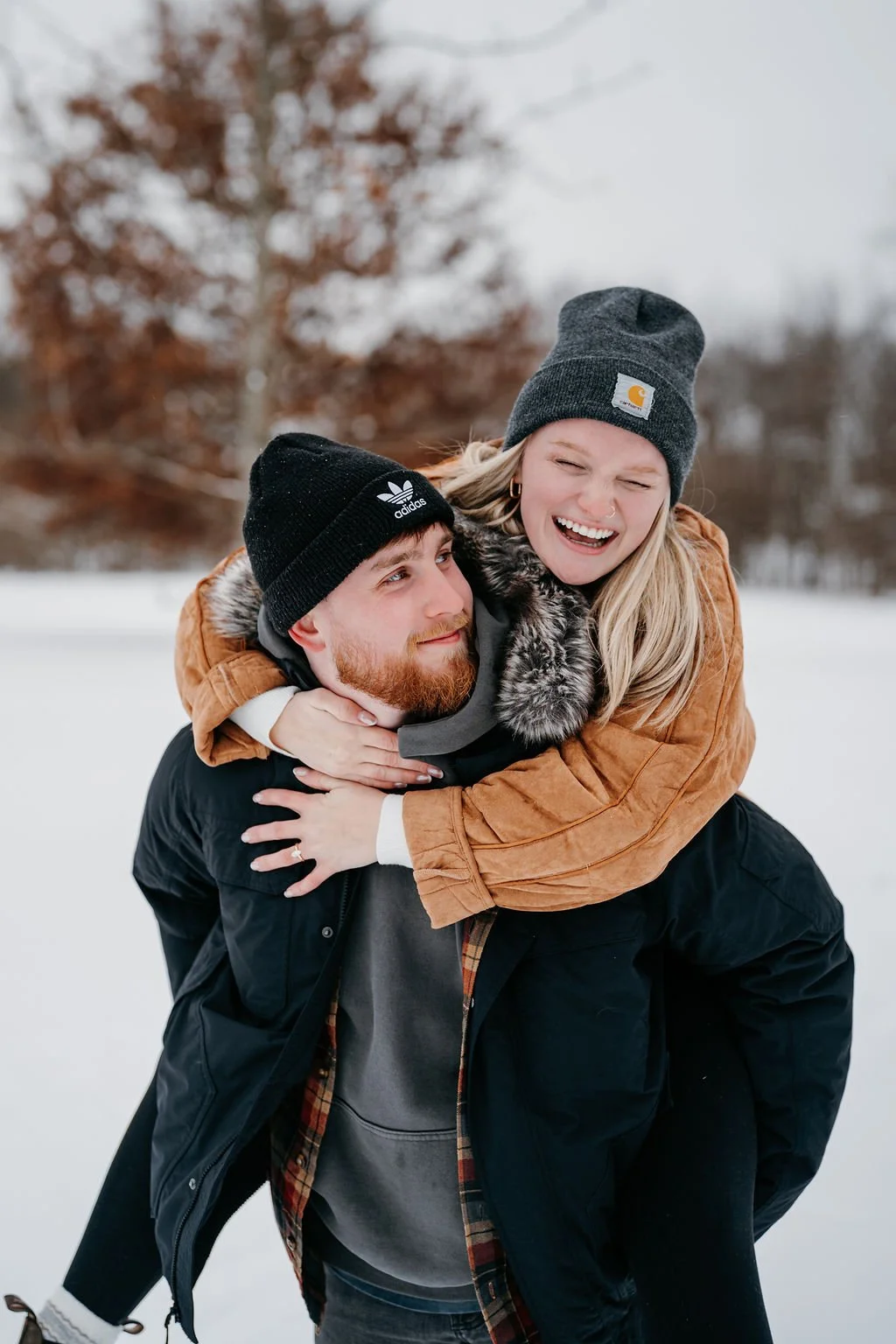 A young couple enjoying a winter day outdoors, with the woman on the man's back, both smiling and dressed warmly with beanies. The background features snow and blurred trees.
