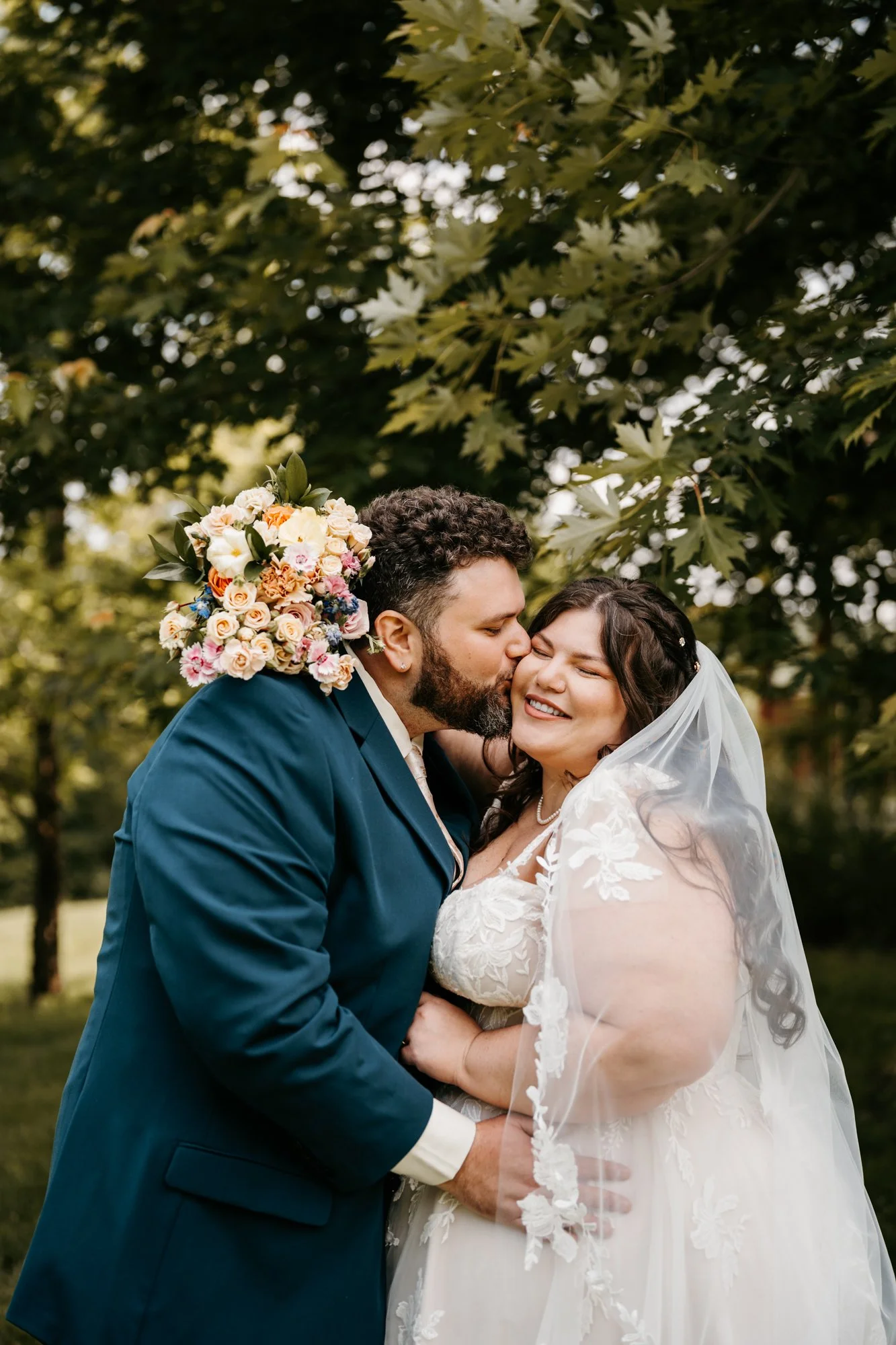A newlywed couple sharing a tender moment outdoors, the groom kissing the bride on the cheek while she smiles, with lush green trees in the background.
