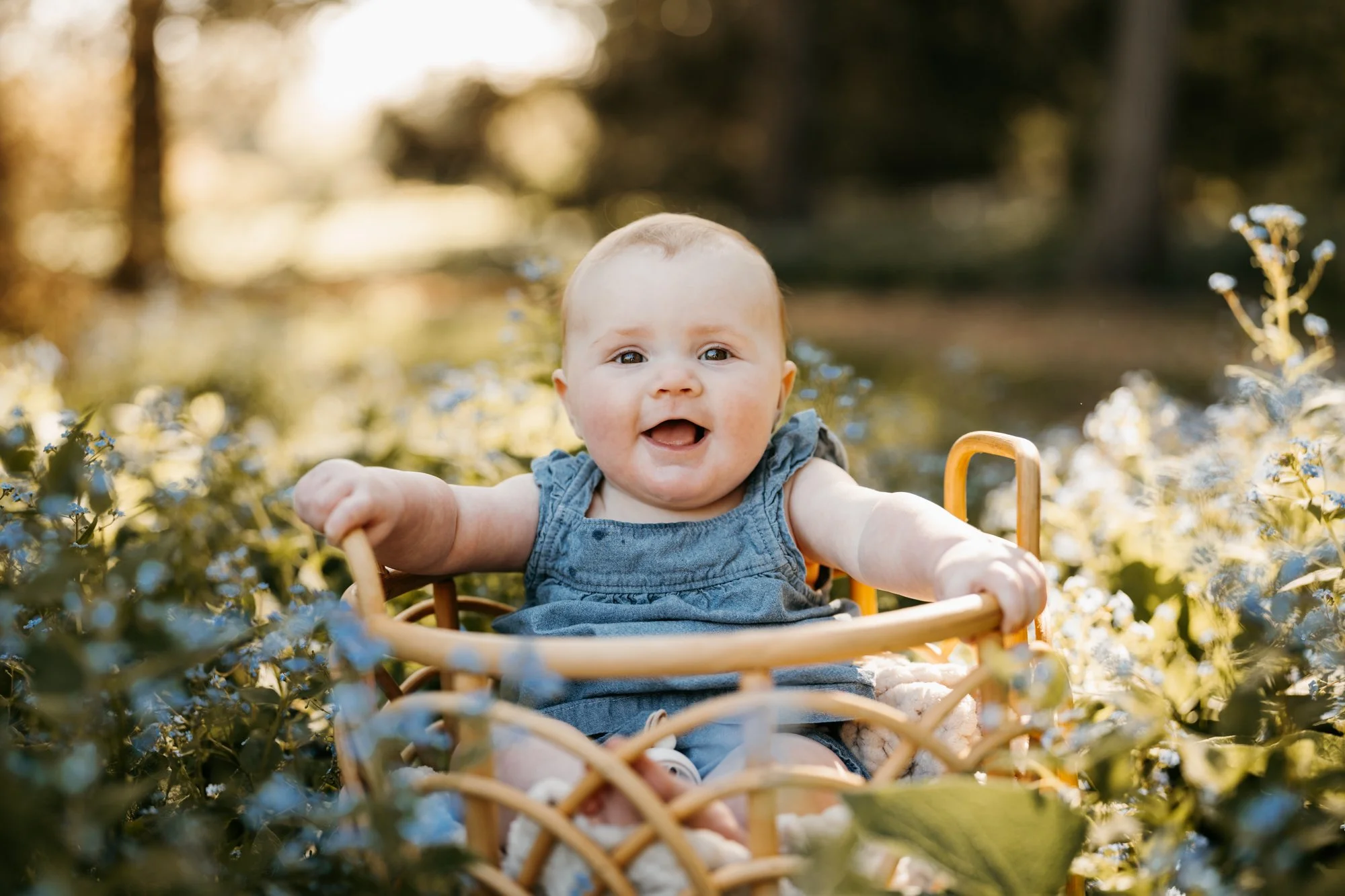 Baby sitting in a wicker basket outdoors surrounded by greenery and flowers, smiling and looking at the camera with a happy expression.
