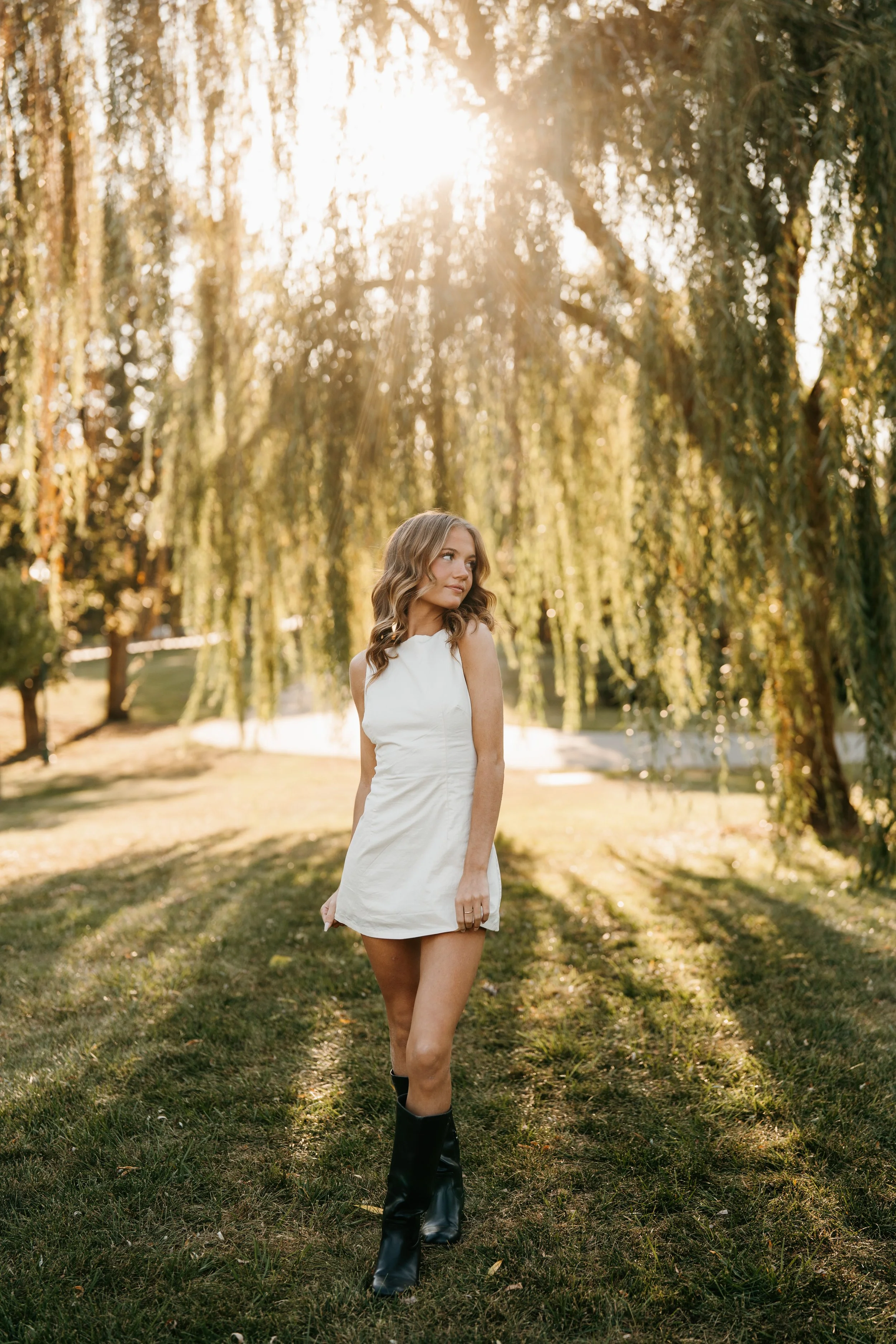 A woman with wavy blonde hair wearing a white sleeveless dress and black boots standing on grass in a park with a large willow tree and sunlight shining through the branches.
