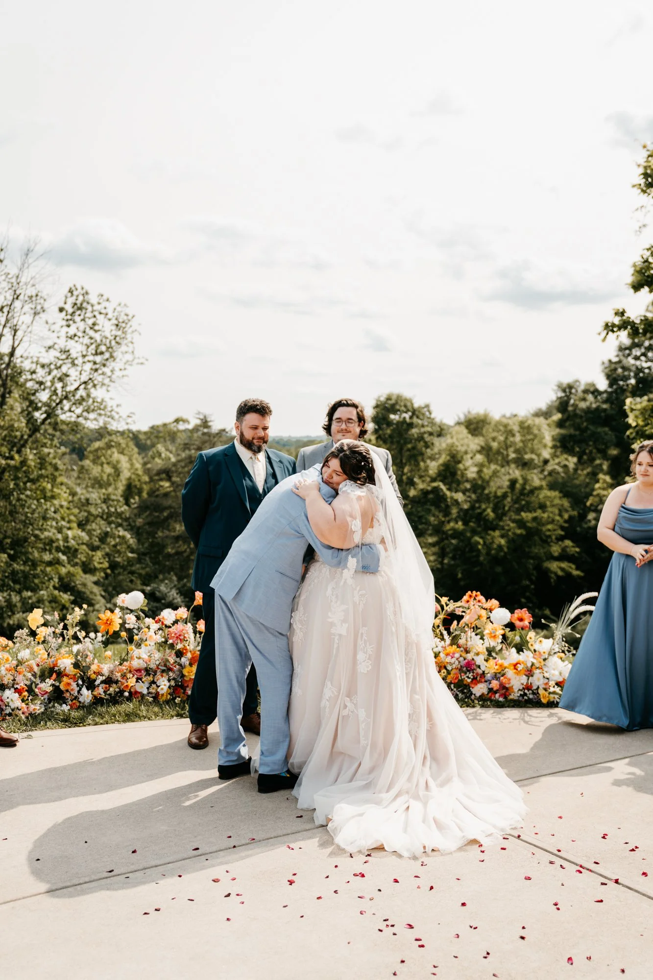 A wedding ceremony outdoors with a bride and groom embracing, surrounded by friends and floral decorations.