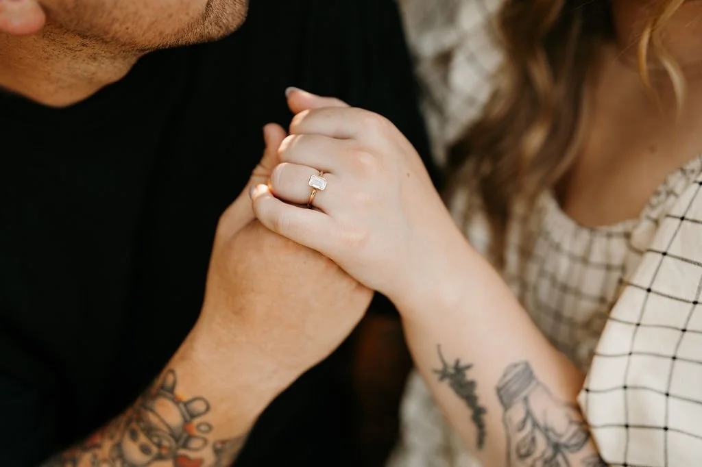 A woman with tattoos on her arm showing her engagement ring as she holds a man's hand, both sitting close together.