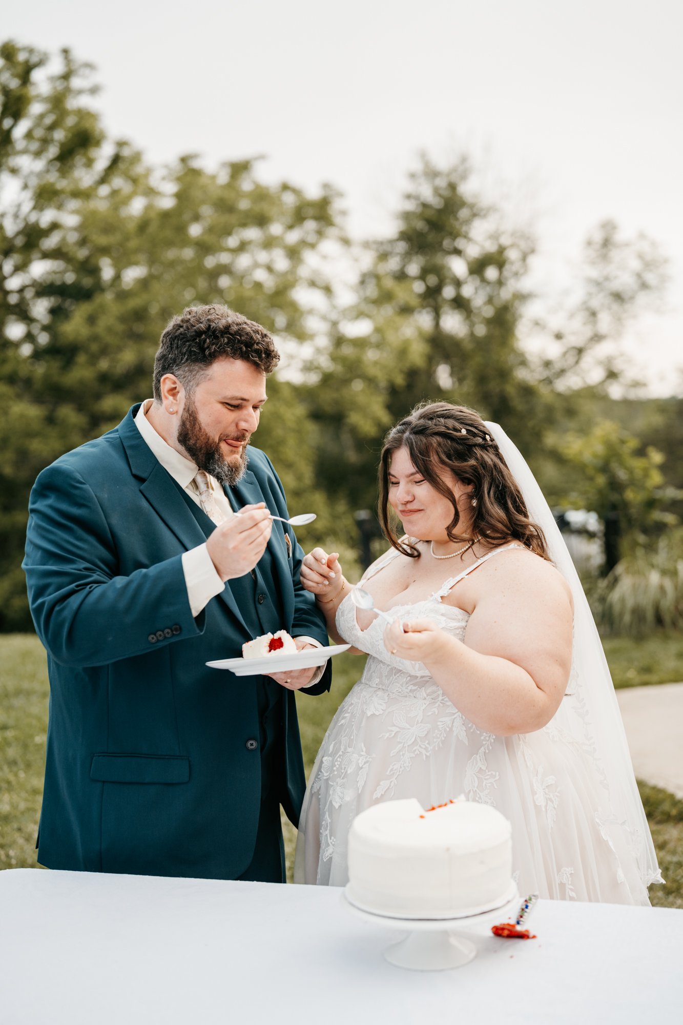 A bride and groom in wedding attire sharing a dessert outdoors at their wedding celebration, with a white cake on the table in front of them.