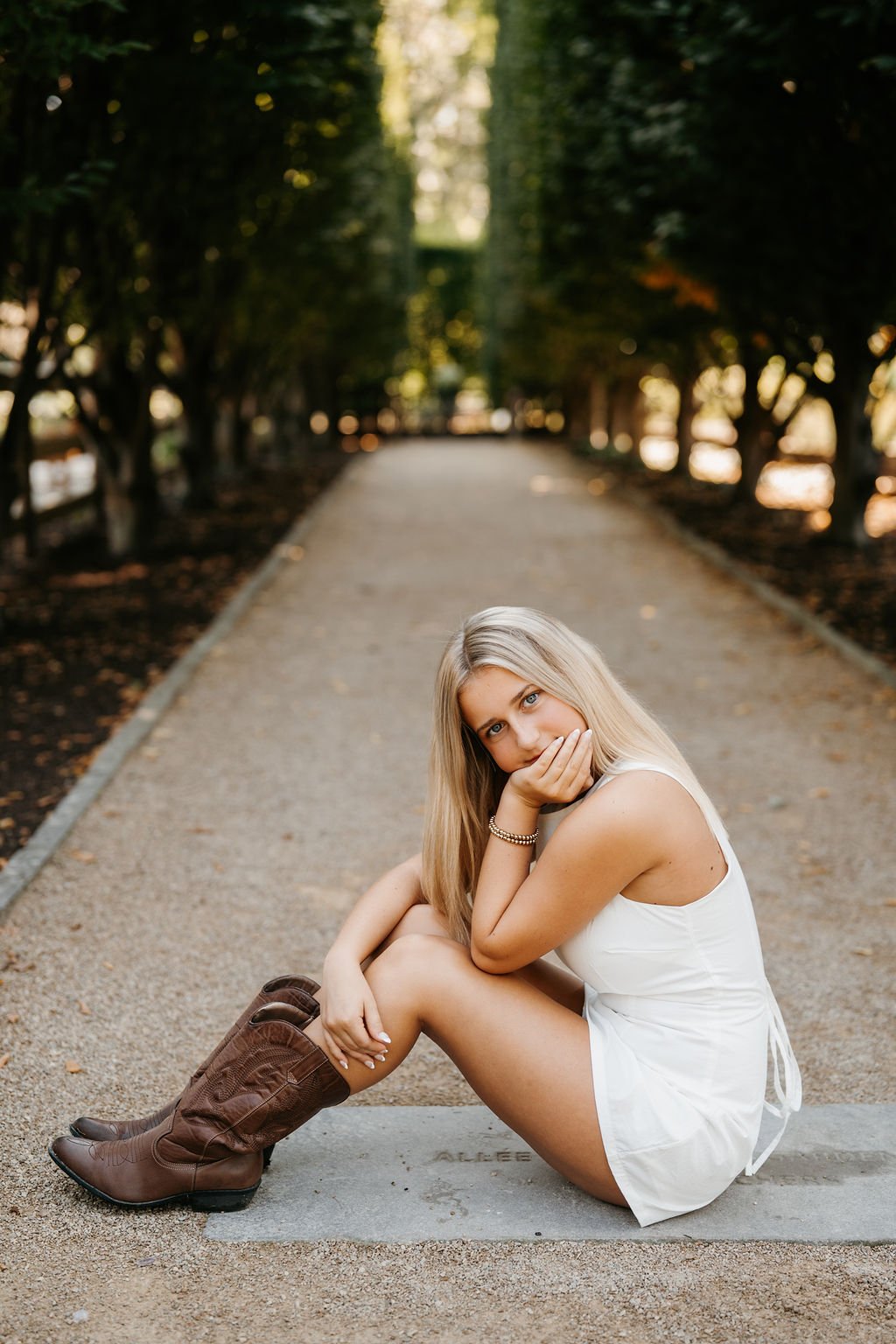 A young woman with long blonde hair sitting on a concrete step on a gravel path, resting her chin on her hand, looking at the camera in a park with trees on both sides.