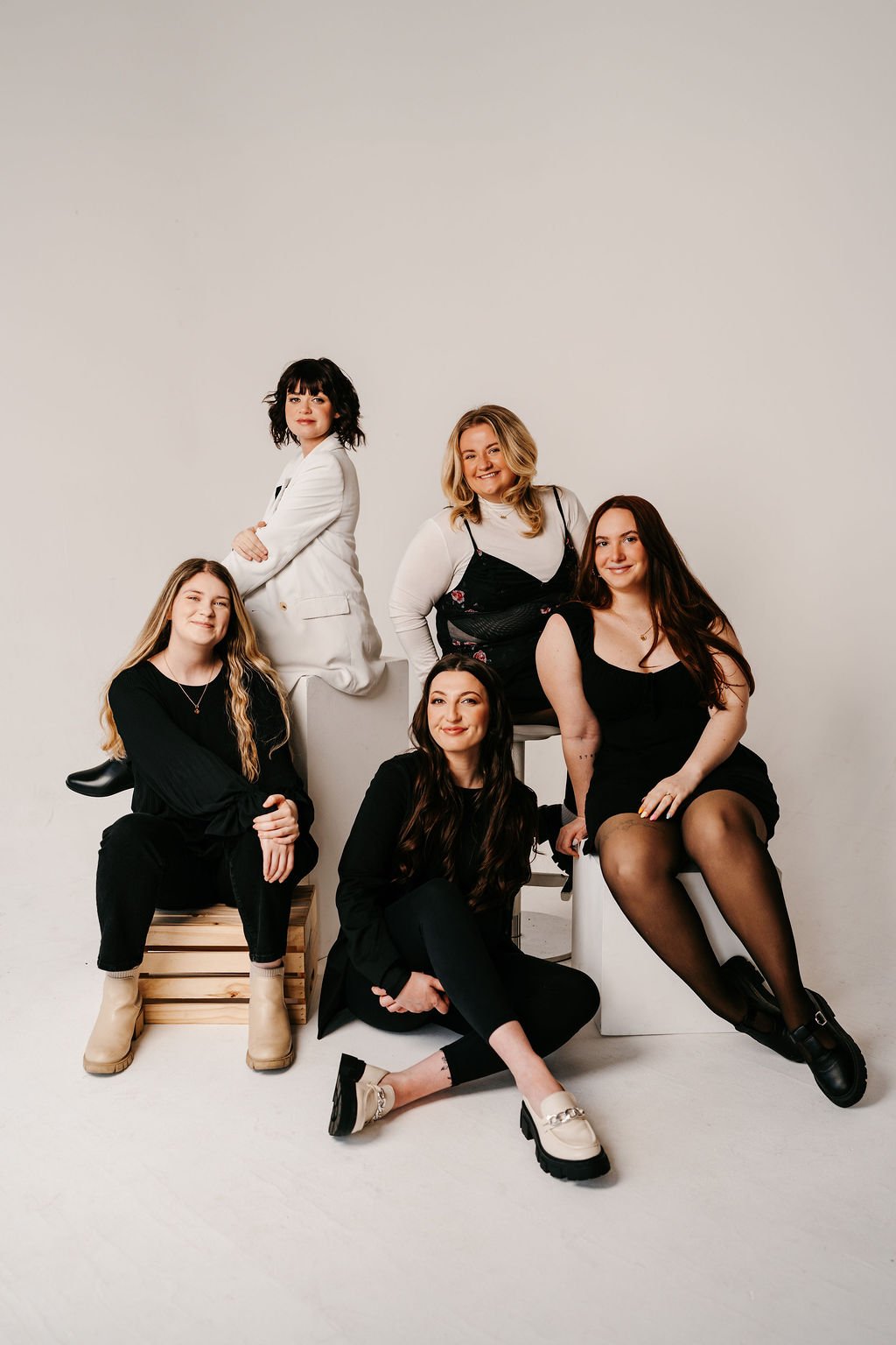 Six women posing in a studio with a plain white background. They are dressed in black and white outfits, with some sitting on stools, one sitting on a wooden crate, and one standing. They are smiling and looking at the camera.