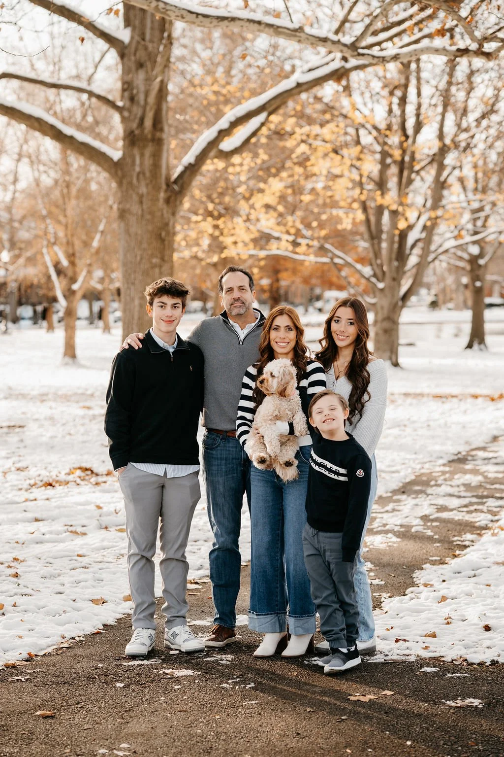 A family of six standing on a snowy park pathway with fall foliage trees in the background, including two adults, two teenagers, two young children, and a dog.