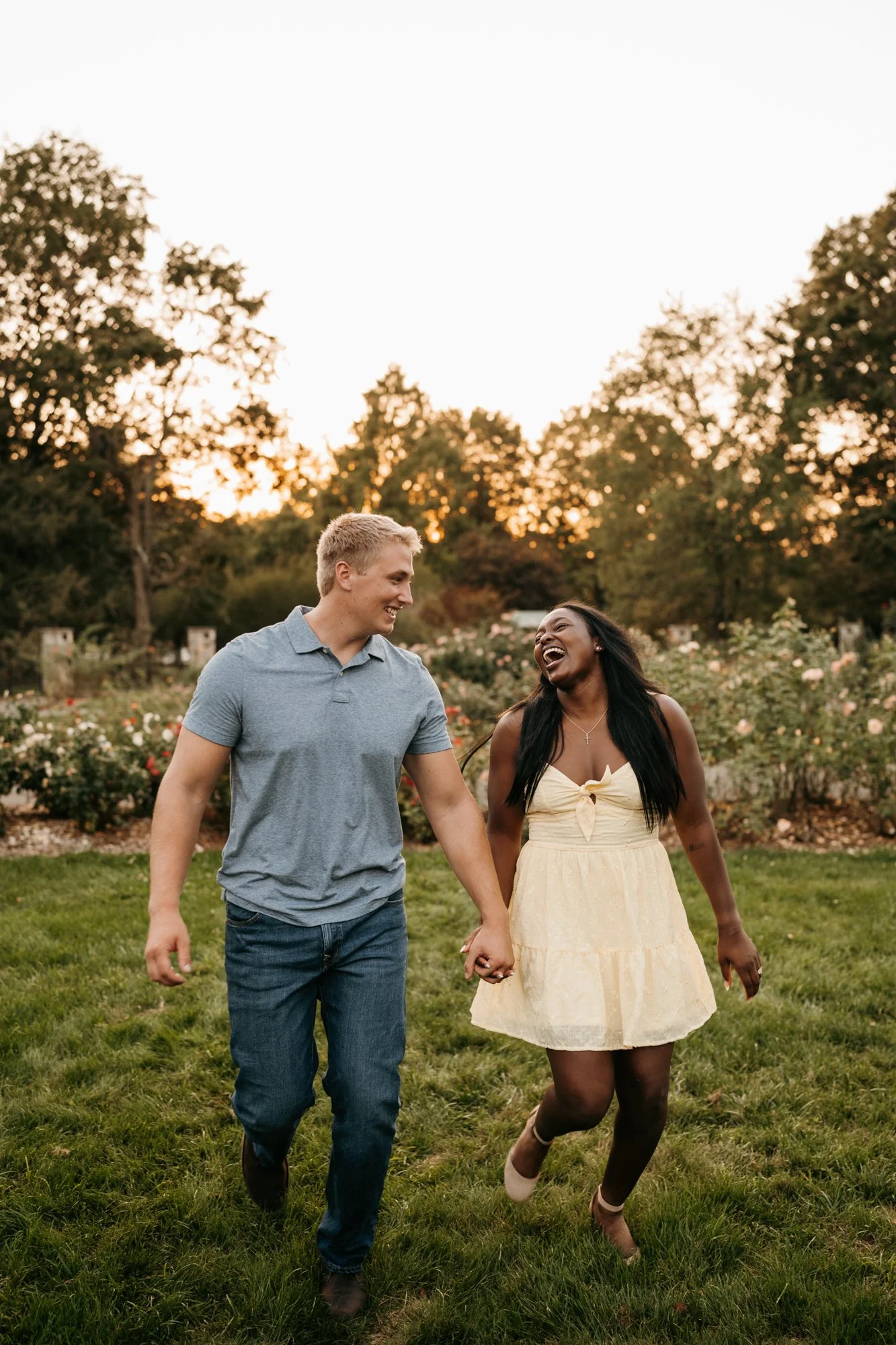 A couple holding hands and walking in a garden at sunset, smiling and laughing.