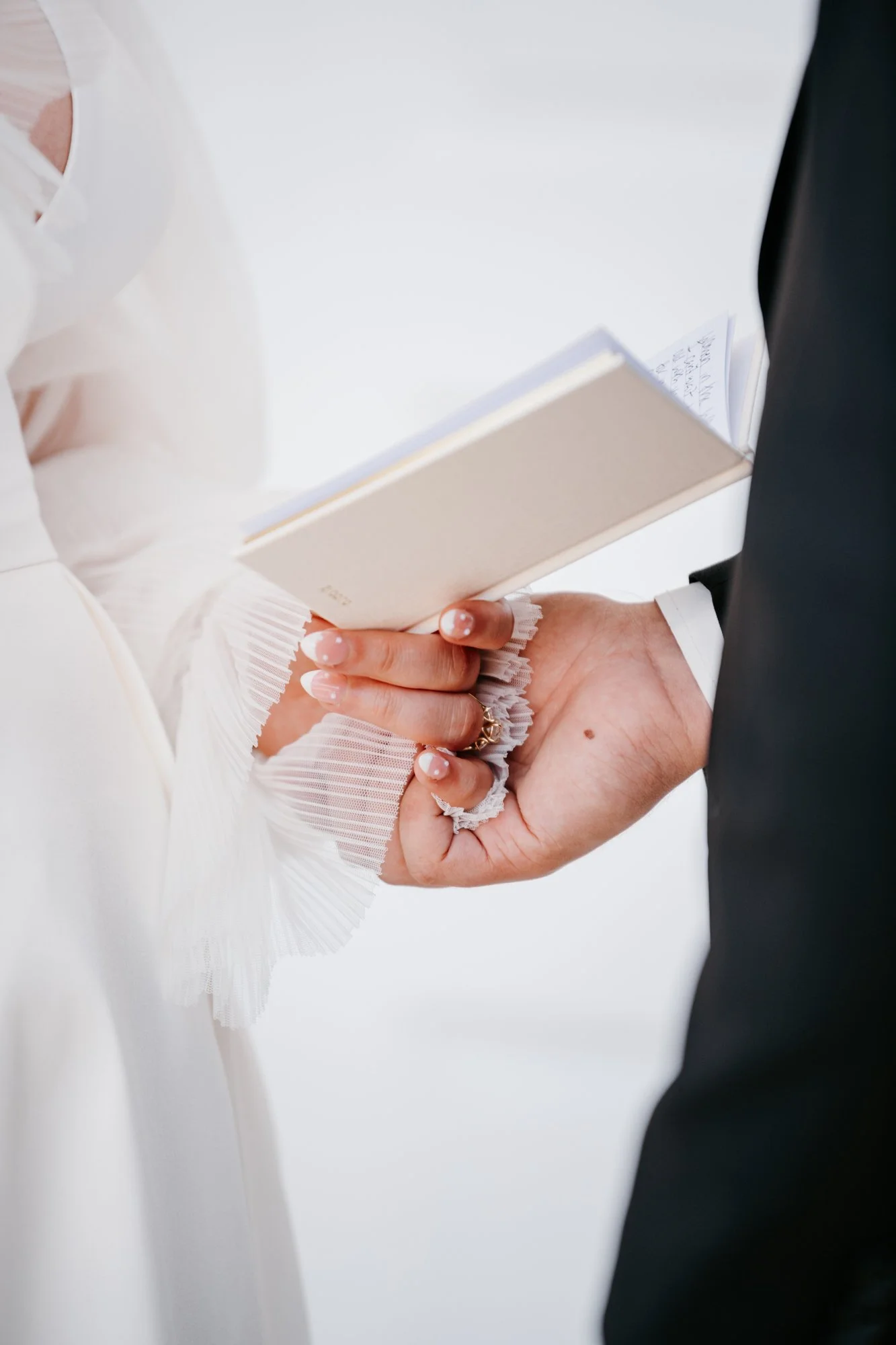 Wedding ceremony with a person holding a small white prayer book, wearing a white dress with pleated cuffs, standing next to another person in a black suit.