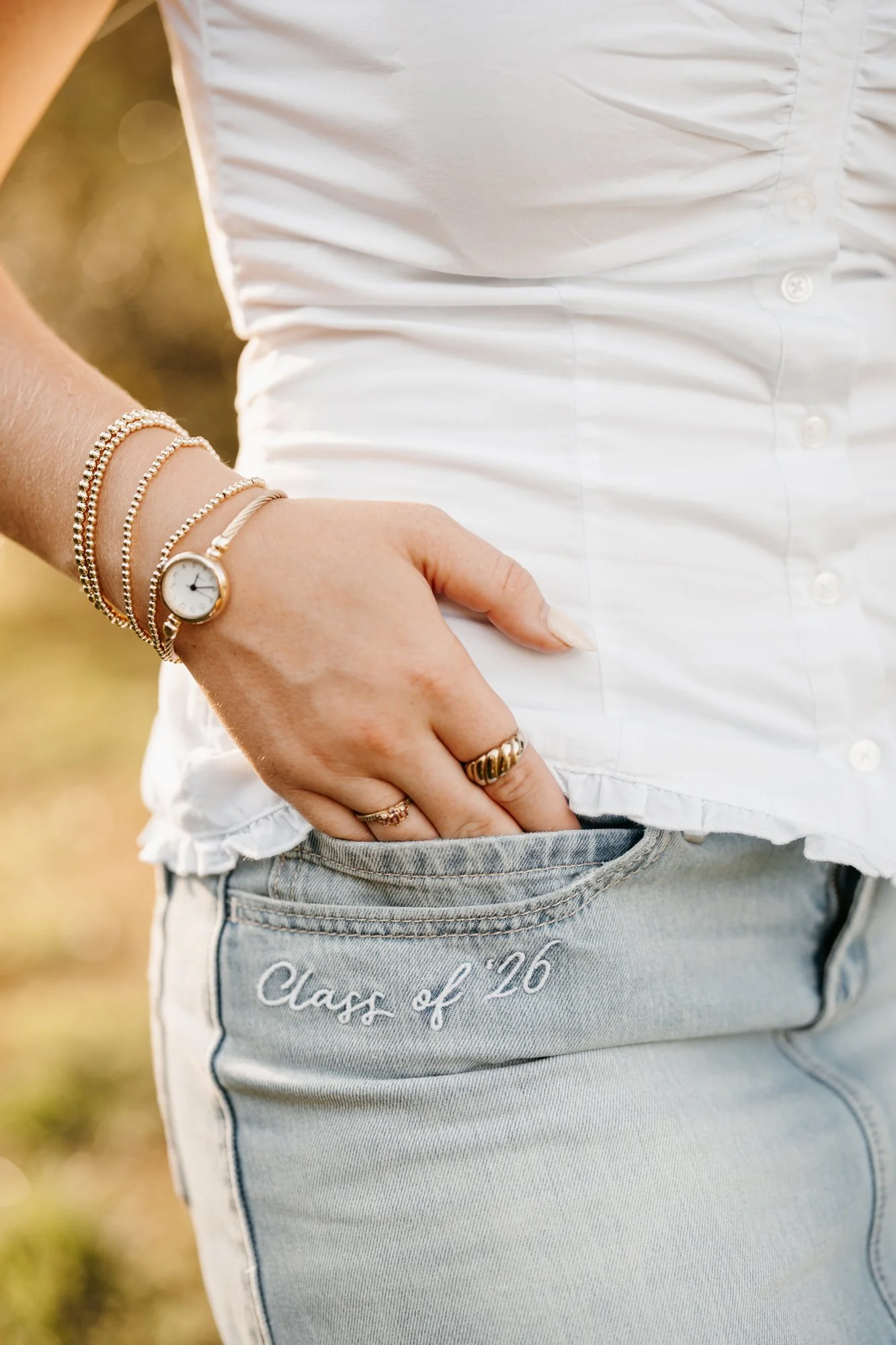 Close-up of a person wearing a white shirt and light-wash jeans with embroidery that says 'Class of '26', standing outdoors with hand in pocket, wearing rings and bracelets.