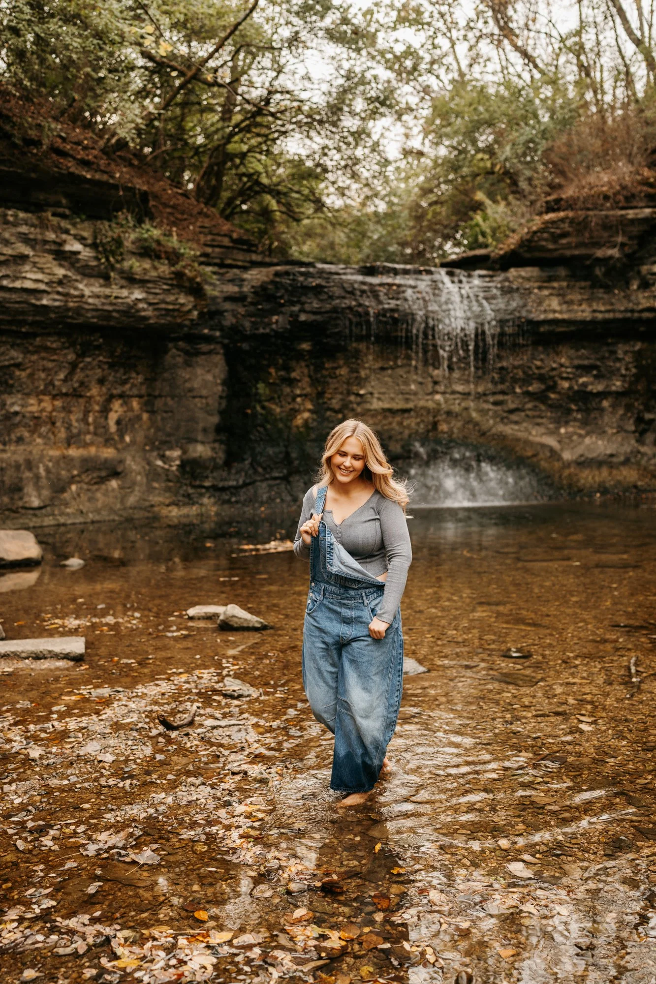 A smiling woman wearing overalls and a gray long-sleeve shirt walking barefoot in a shallow creek with a waterfall and trees in the background.