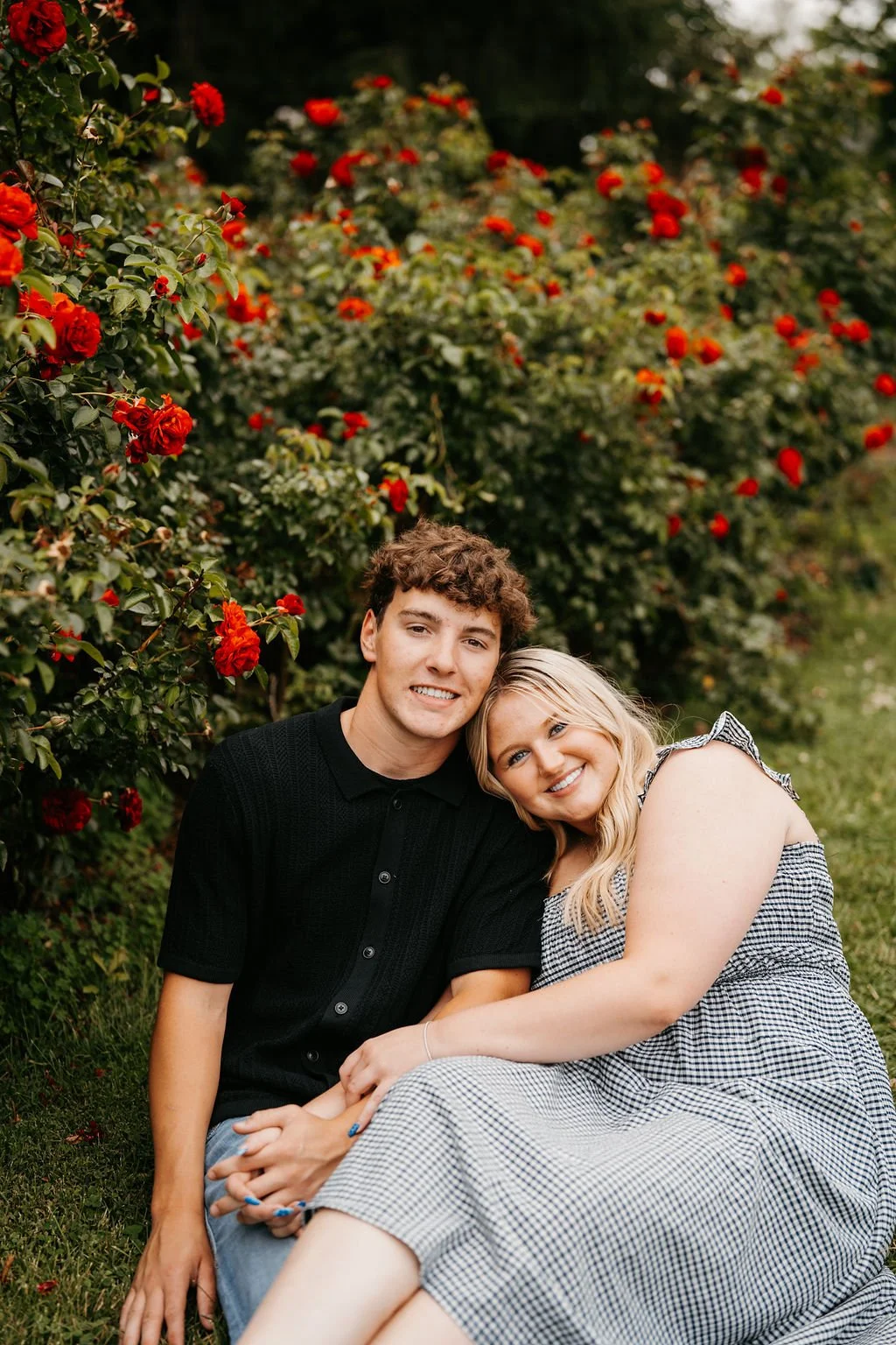 A young man and woman sitting on the grass in front of red flowering bushes, smiling and leaning their heads together.