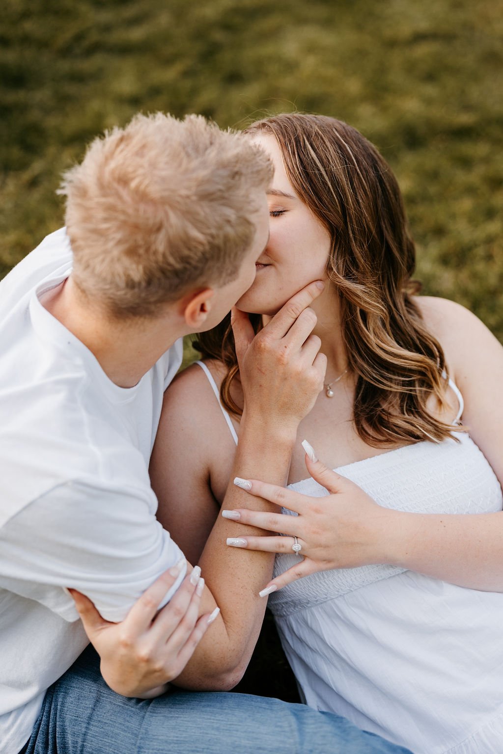 A couple sharing a kiss outdoors, with the woman touching the man's chin and holding his arm, greenery in the background.