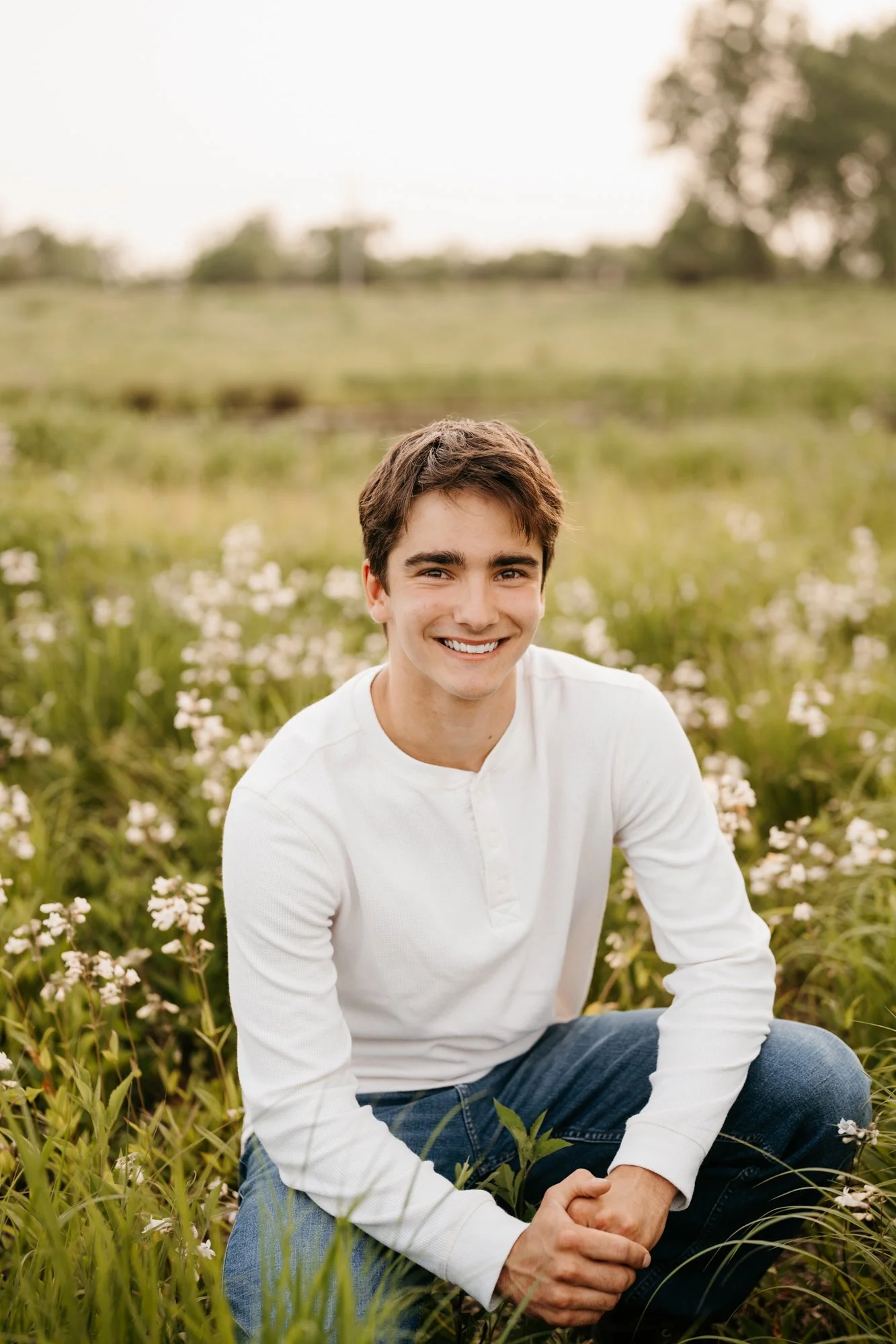 A young man with dark hair, wearing a white long sleeve shirt and blue jeans, smiling while sitting in a green grassy field with small white flowers, during daytime with overcast sky.