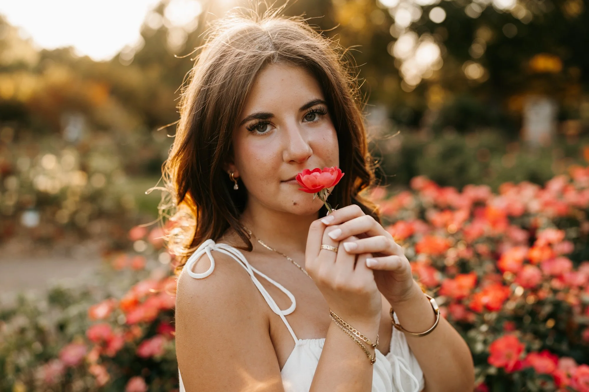A young woman with wavy brown hair and jewelry holds a red flower near her face in a garden with blooming roses at sunset.