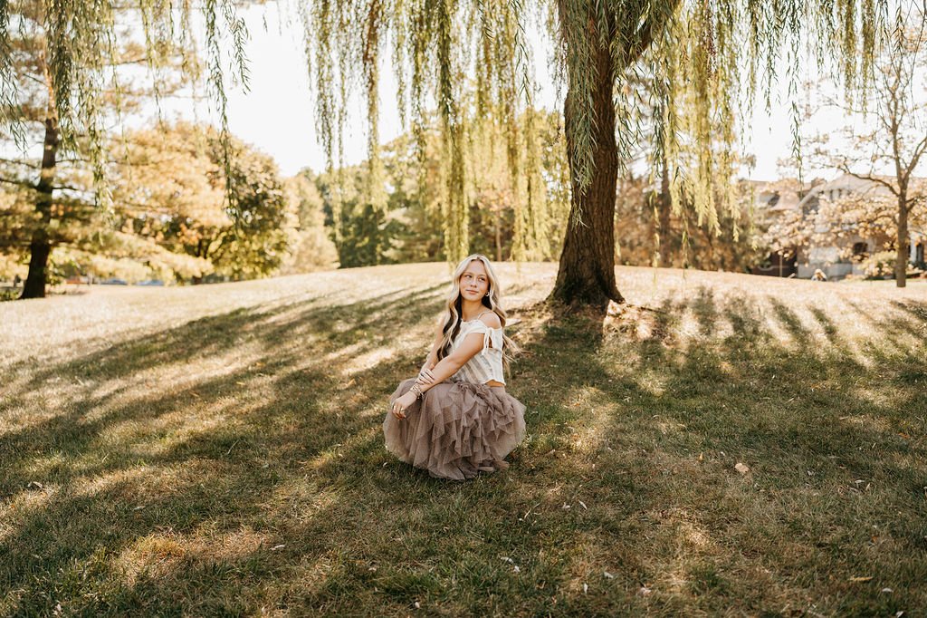 A young girl in a white top and brown tulle skirt sitting under a large tree with hanging branches in a park during autumn, with fallen leaves and a residential neighborhood in the background.