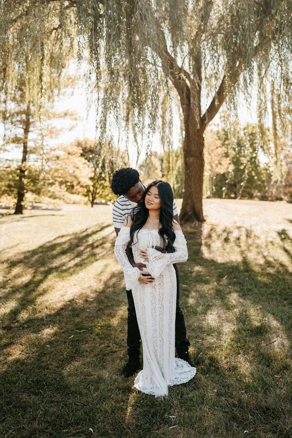 A couple stands under a large tree in a park during fall, with the man hugging the woman from behind. The woman wears a white lace dress, and the man wears a white and black striped shirt. They appear happy and content.