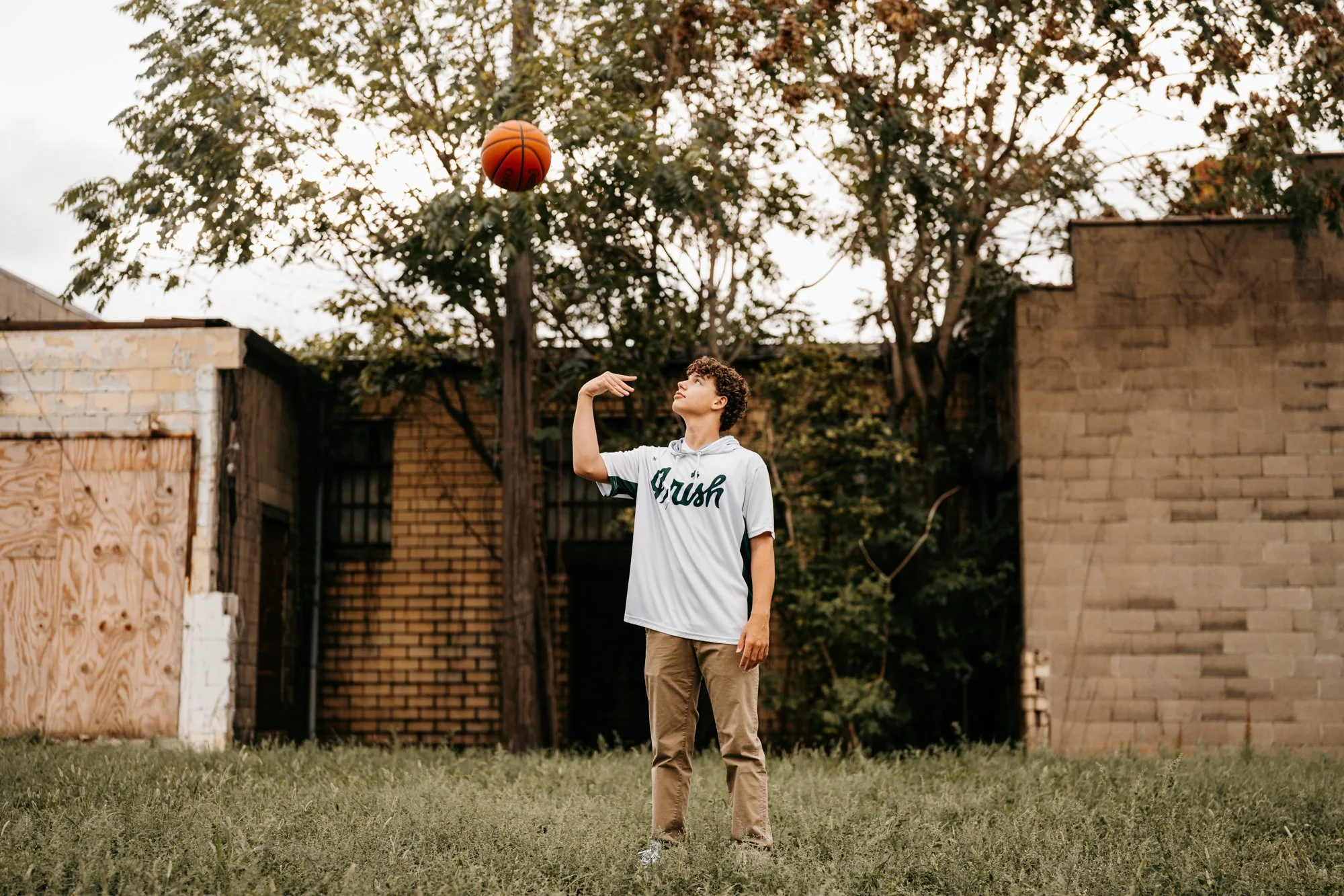 A young man playing basketball outdoors on a grassy area, wearing a white jersey with the word 'Fresh' on it, and looking up at a basketball in the air.
