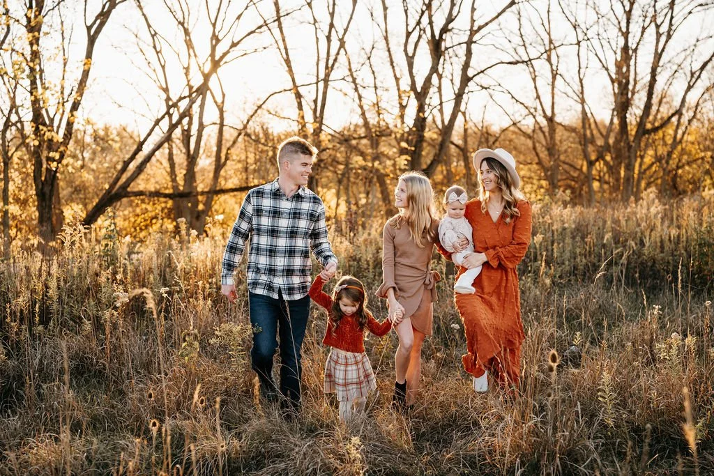Family walking through a field of tall grass during sunset, with leafless trees in the background. The family includes a man, a woman, and three young girls, all dressed in fall clothing.