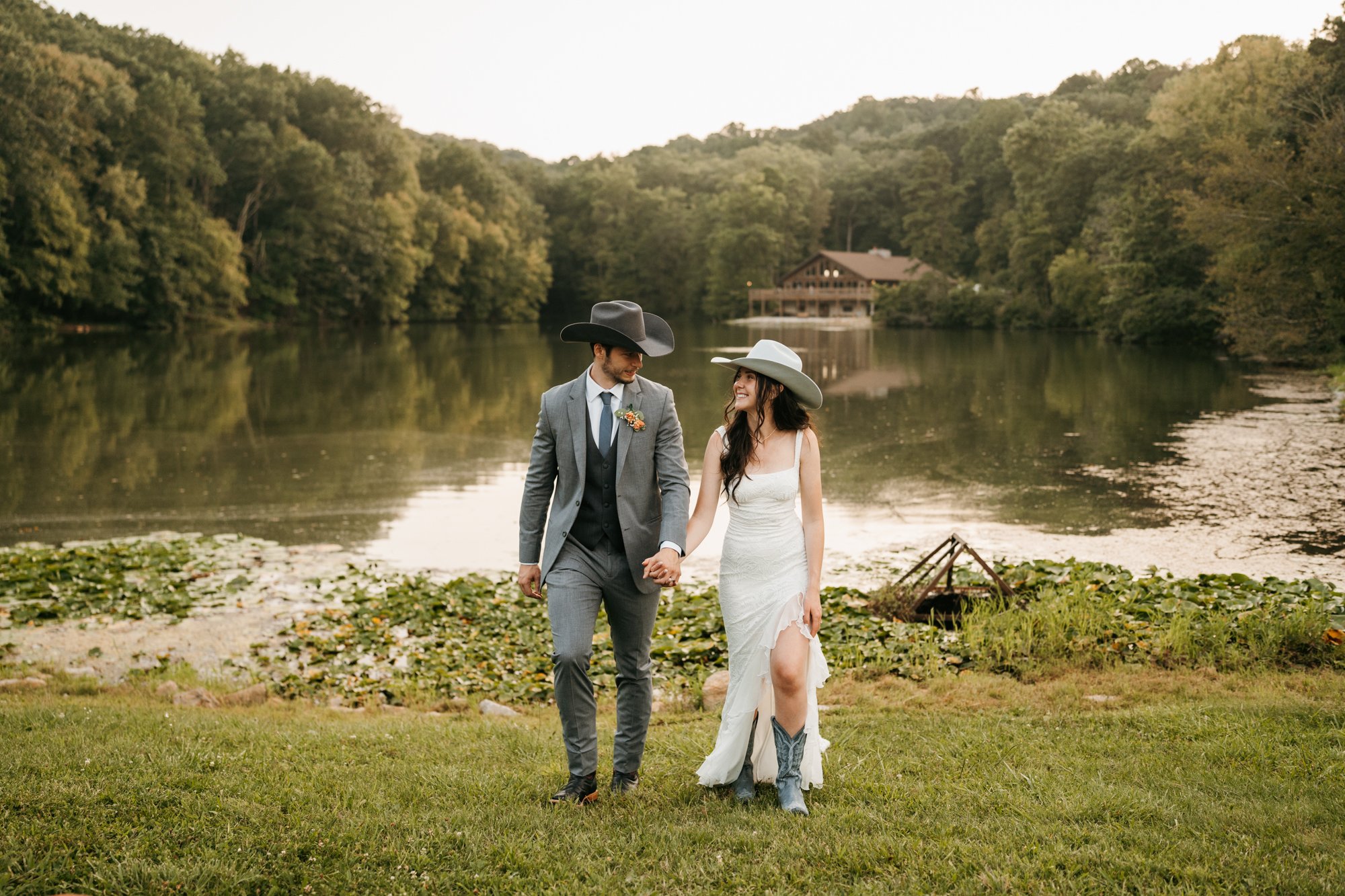 A couple dressed in wedding attire holding hands, walking on a grassy area near a lake, surrounded by trees, with a wooden house in the background.