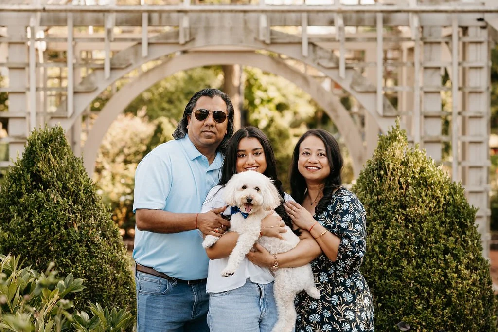 A family of three with their dog standing outdoors in front of a wooden arch and greenery, smiling at the camera.