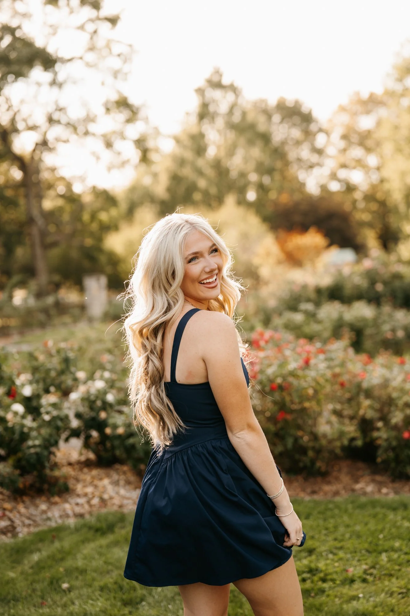 A young woman with long blonde hair, smiling and looking back over her shoulder, standing outdoors in a garden with blooming flowers and trees, during sunset.