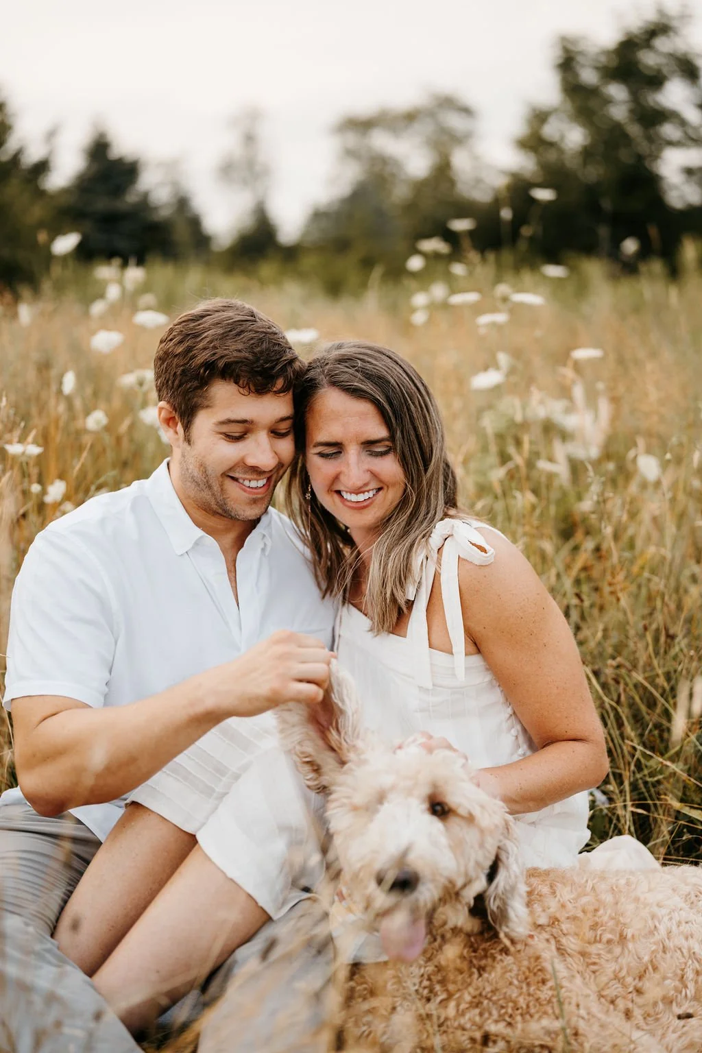 A happy couple sitting in a field of tall grass and wildflowers with a fluffy dog, smiling and playing with the dog’s paw.