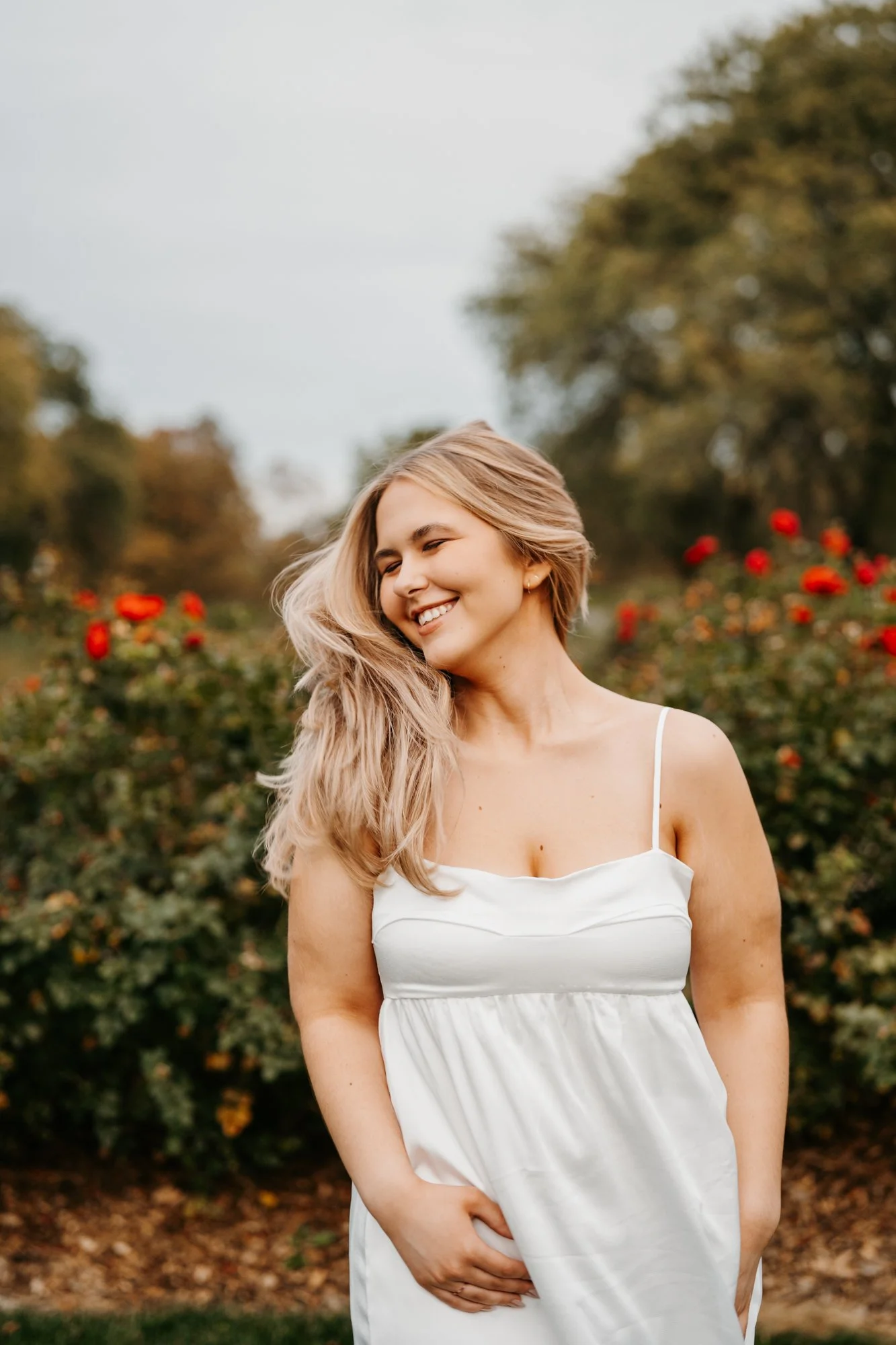 Smiling woman in a white dress standing outdoors among red flowers and green trees on an overcast day.